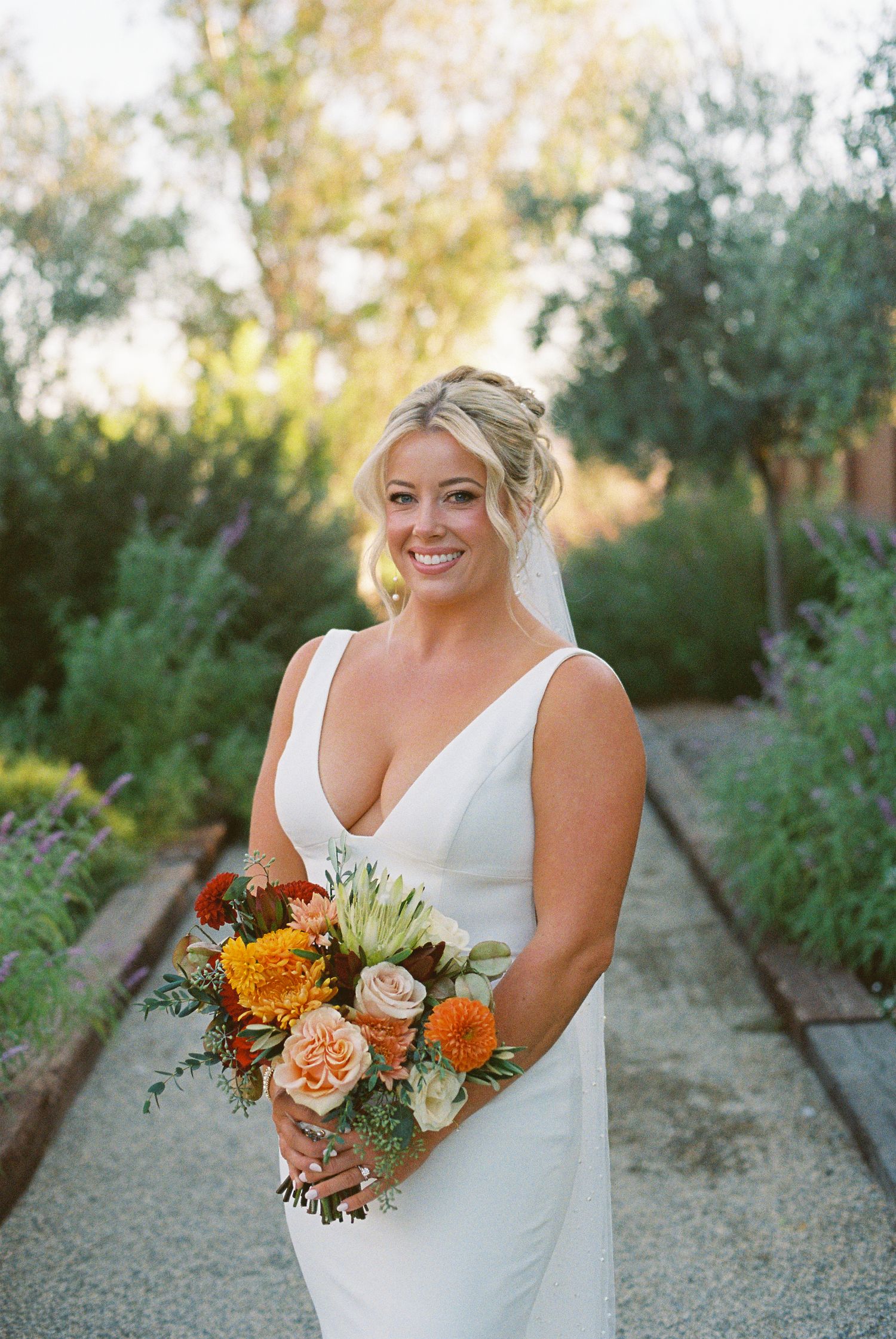 Smiling bride in white V-neck gown holding vibrant orange and peach bouquet, standing in lush garden path at golden hour.