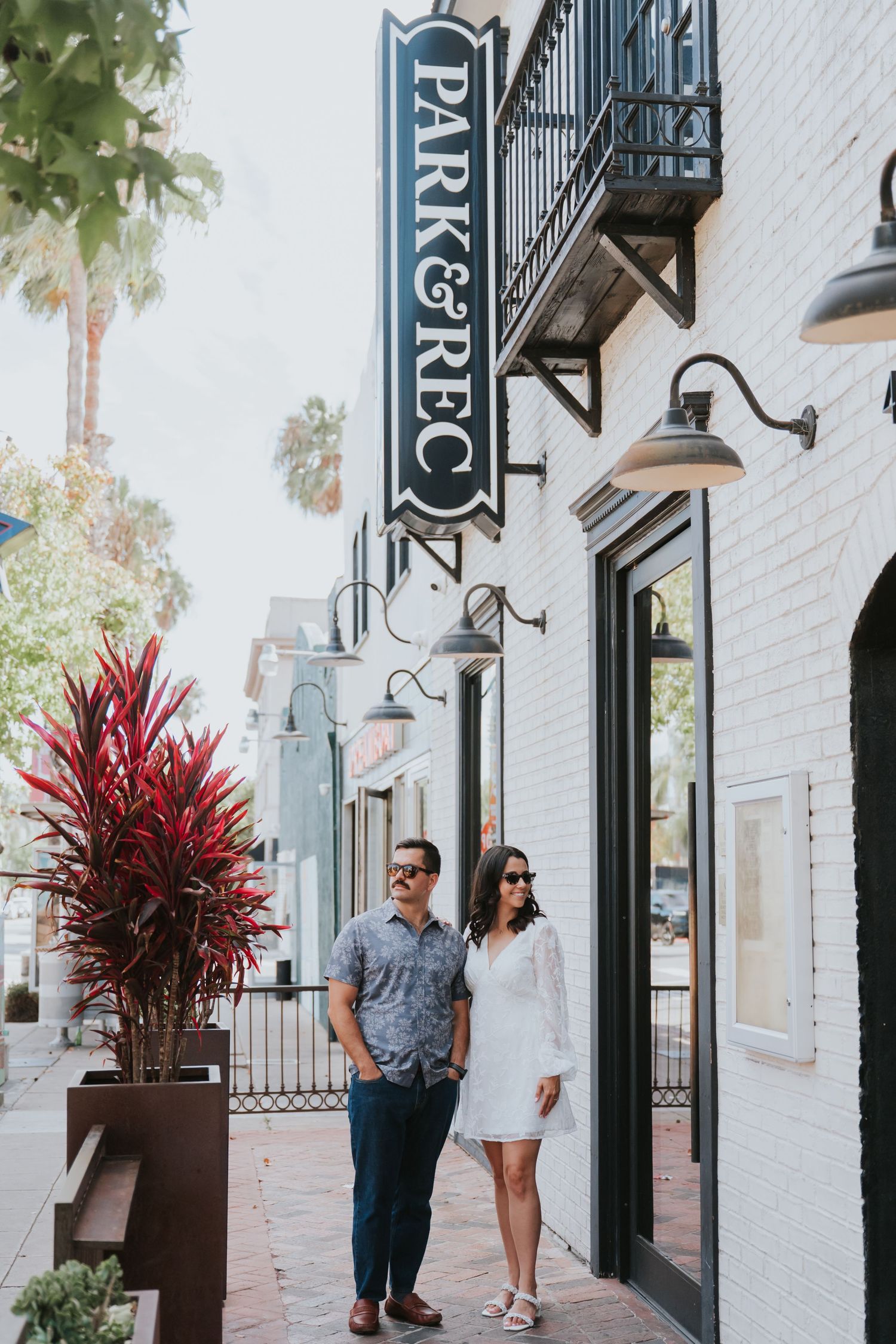 A couple walks along a white building exterior with black trim and decorative red plants in modern planters.