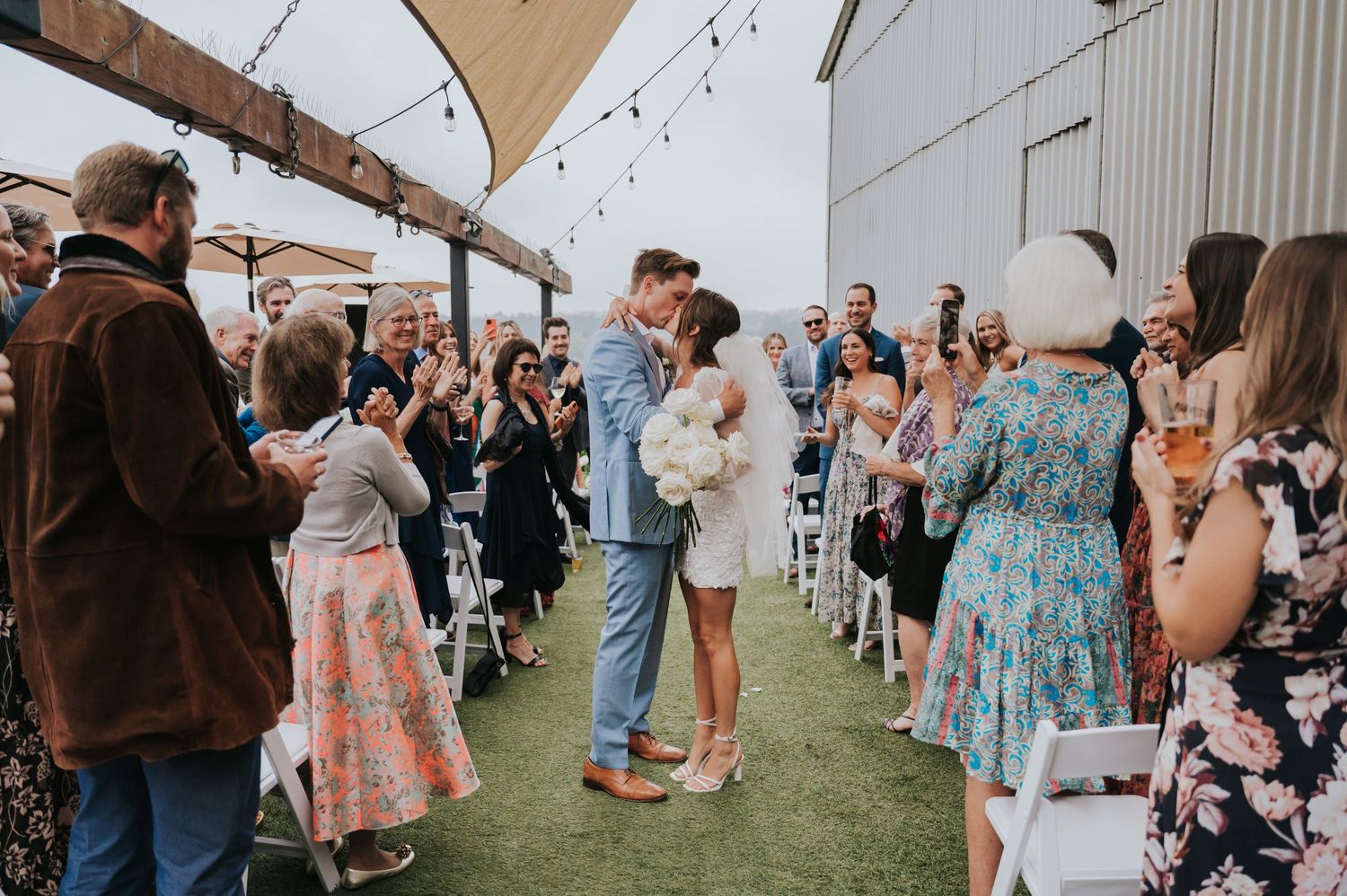 Wedding guests gather outdoors under string lights for a celebration at a rustic barn venue.