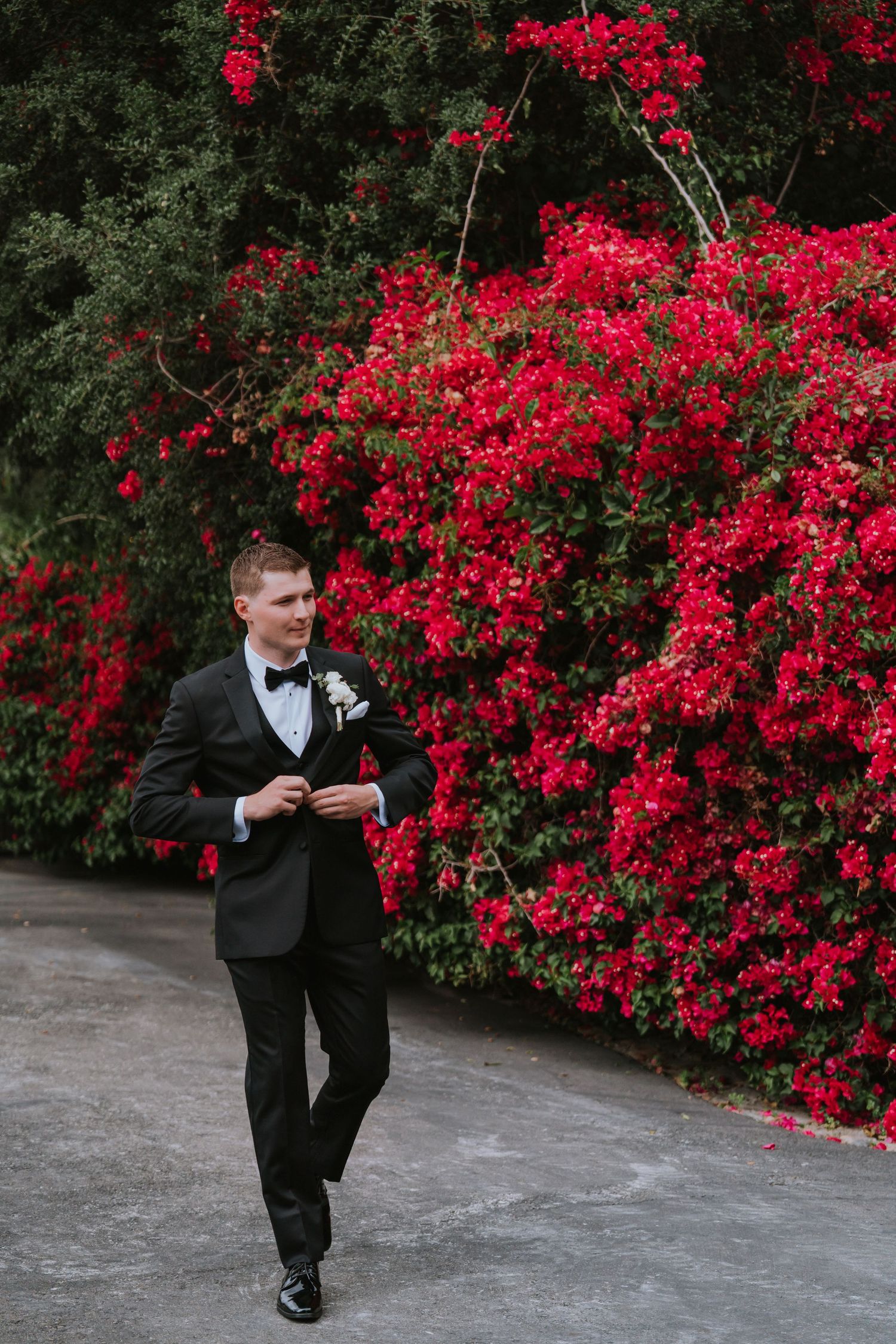 A person in formal attire poses near blooming red bougainvillea bushes.