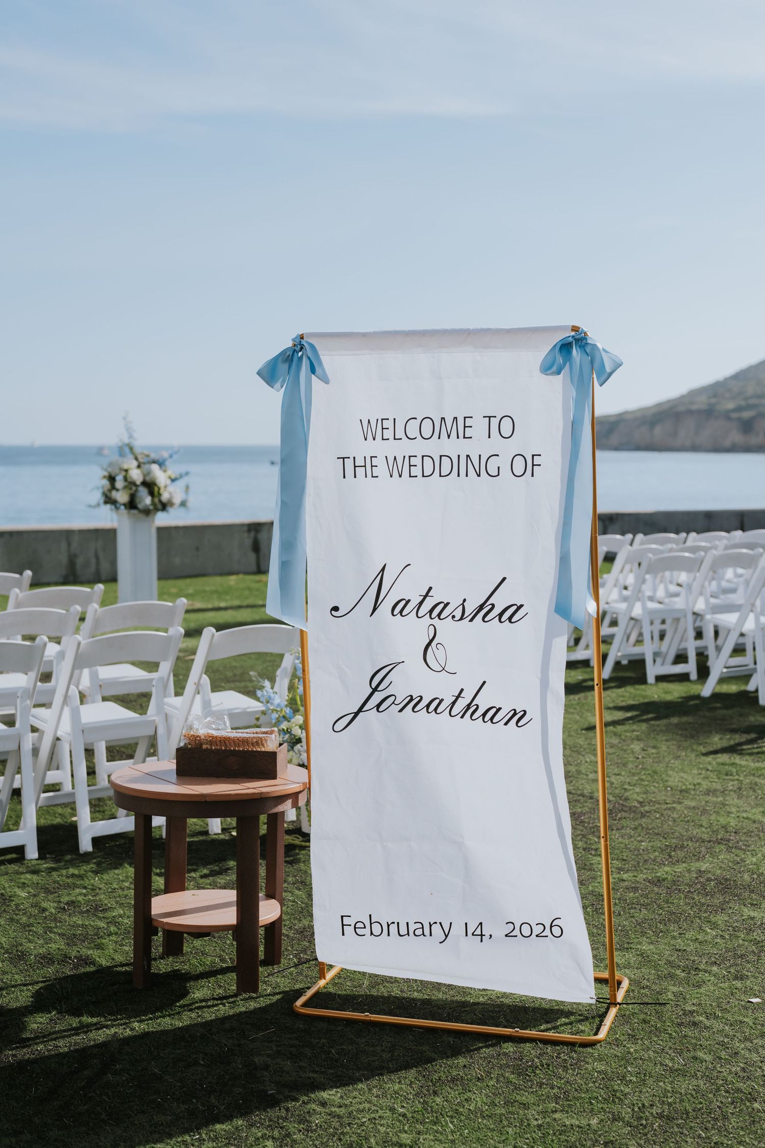 Outdoor wedding welcome sign for Natasha and Jonathan on February 14, 2026, with blue ribbon bows and ocean backdrop.