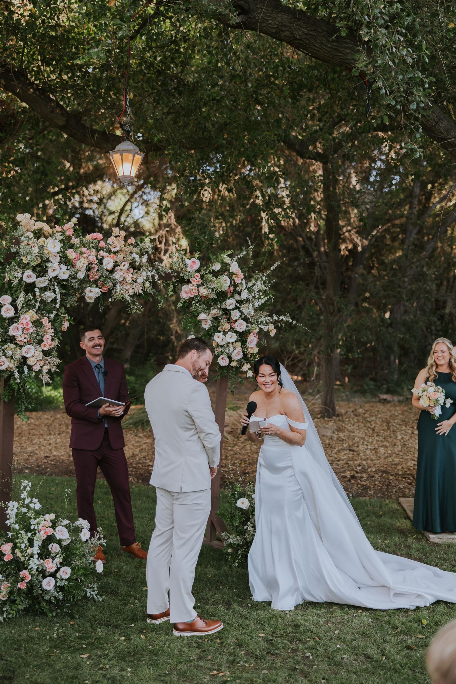 Bride reads vows to groom at outdoor wedding ceremony with pink floral arch and officiant in burgundy suit.