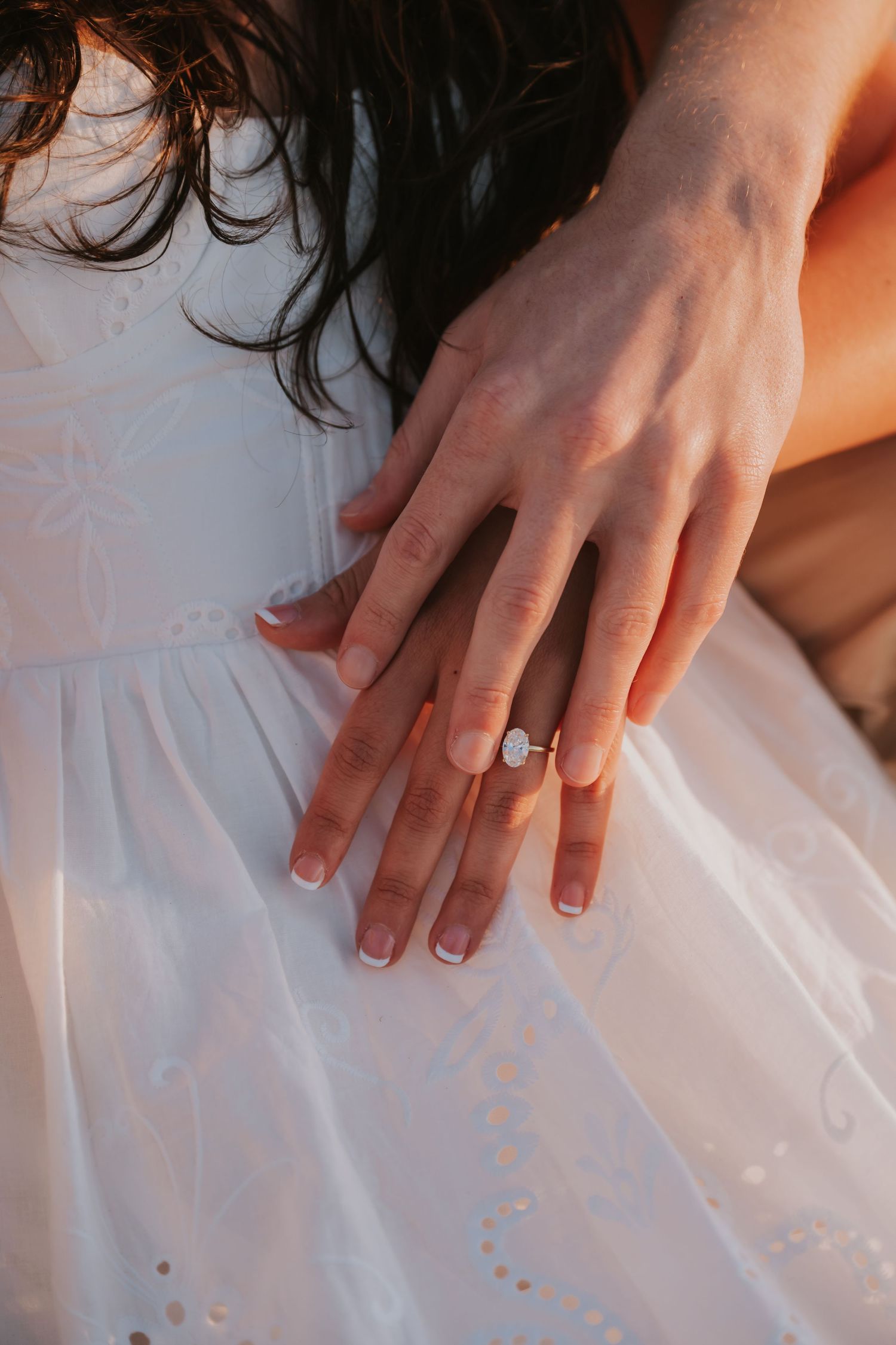 Couple showing off engagement ring with diamond solitaire, hands resting on white eyelet dress in warm golden light.