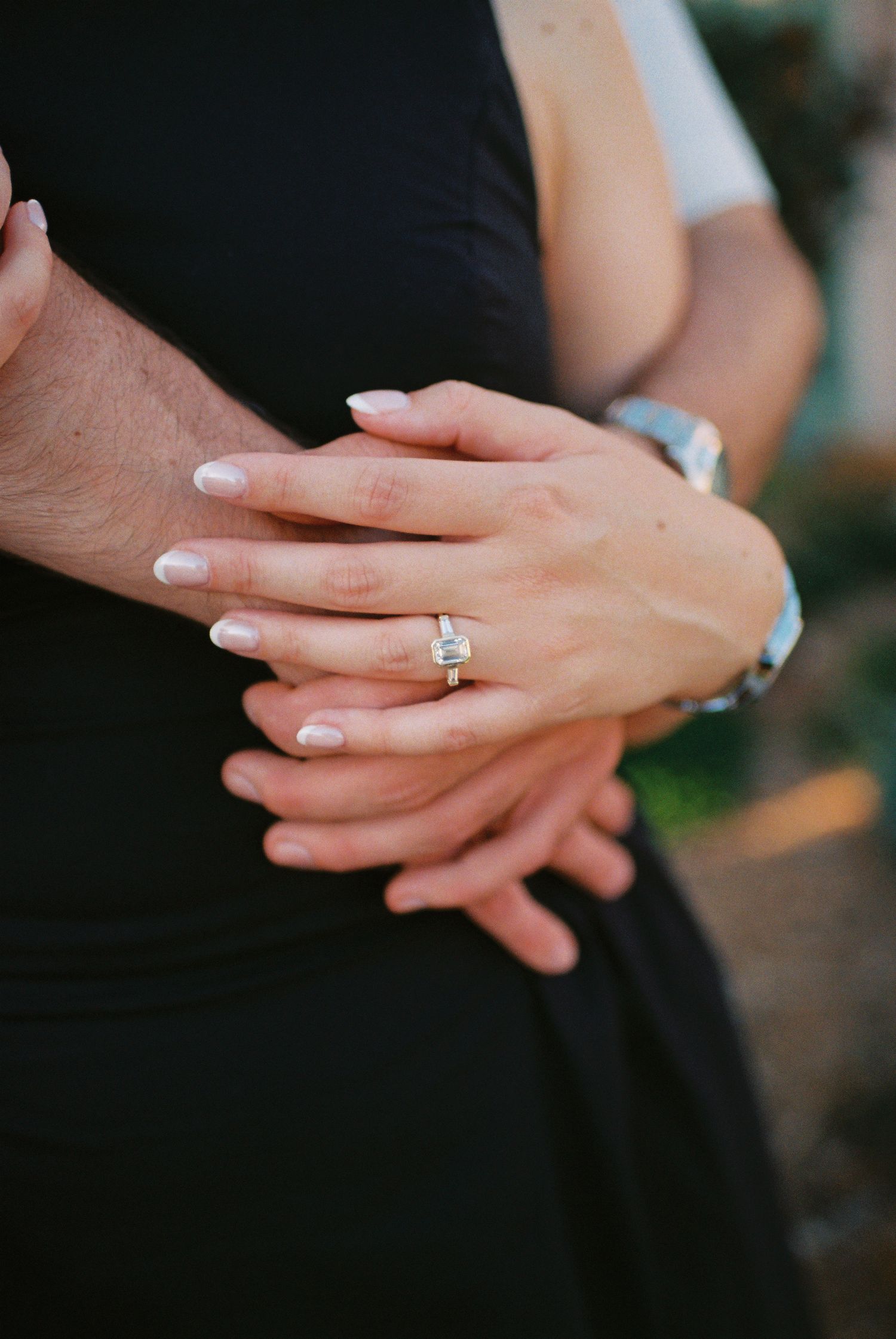 Close-up of couple's hands embracing, showcasing a stunning emerald-cut diamond engagement ring on woman's finger.