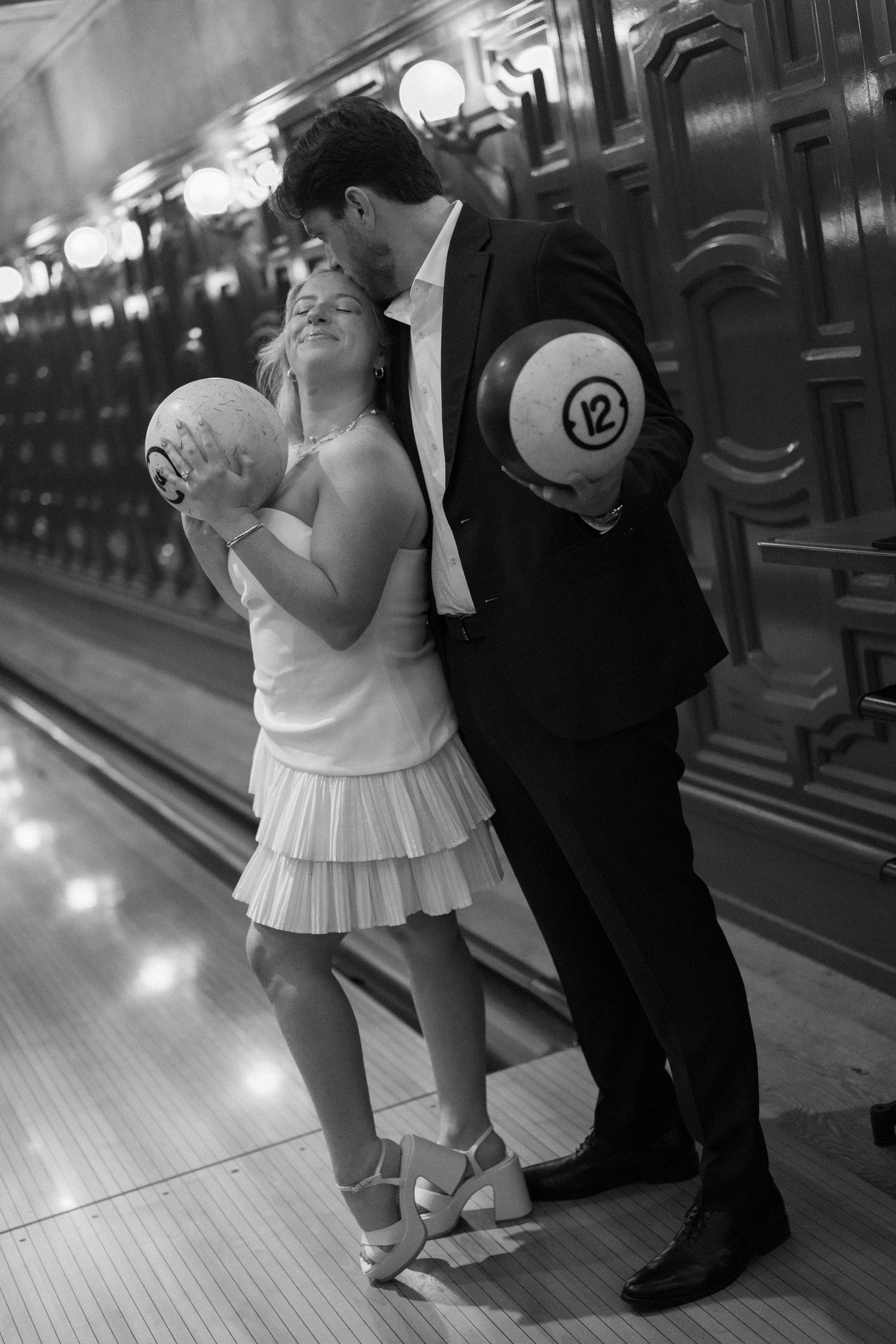 Black and white photo of couple holding bowling balls at a vintage bowling alley, sharing a kiss.