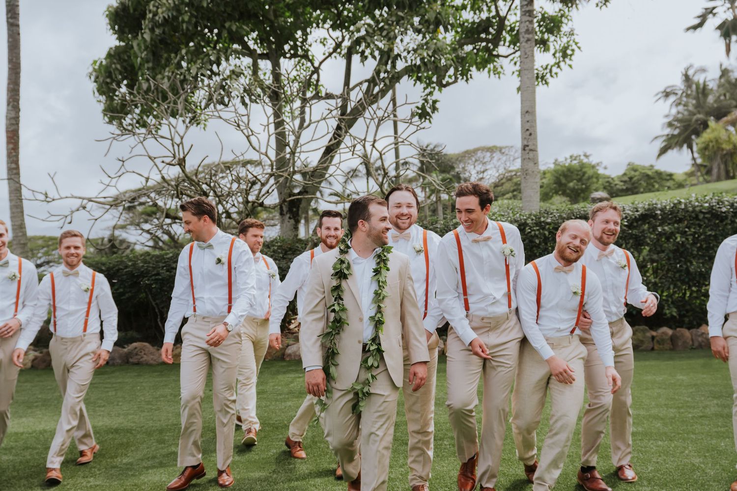Groomsmen in khaki pants and suspenders pose together on a tropical lawn.