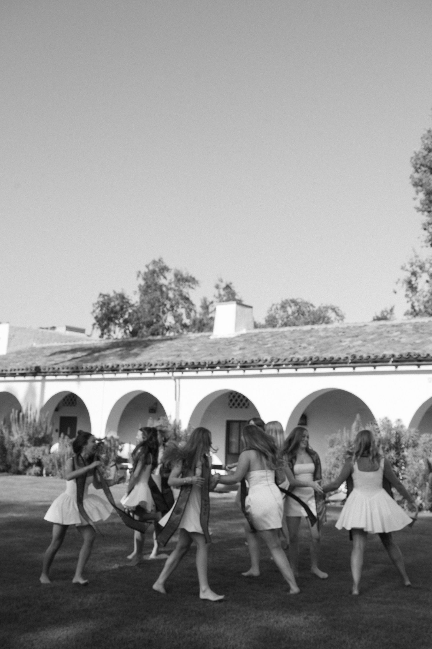 Black and white photo of bridesmaids in white dresses dancing in a circle outdoors near a Spanish-style building.