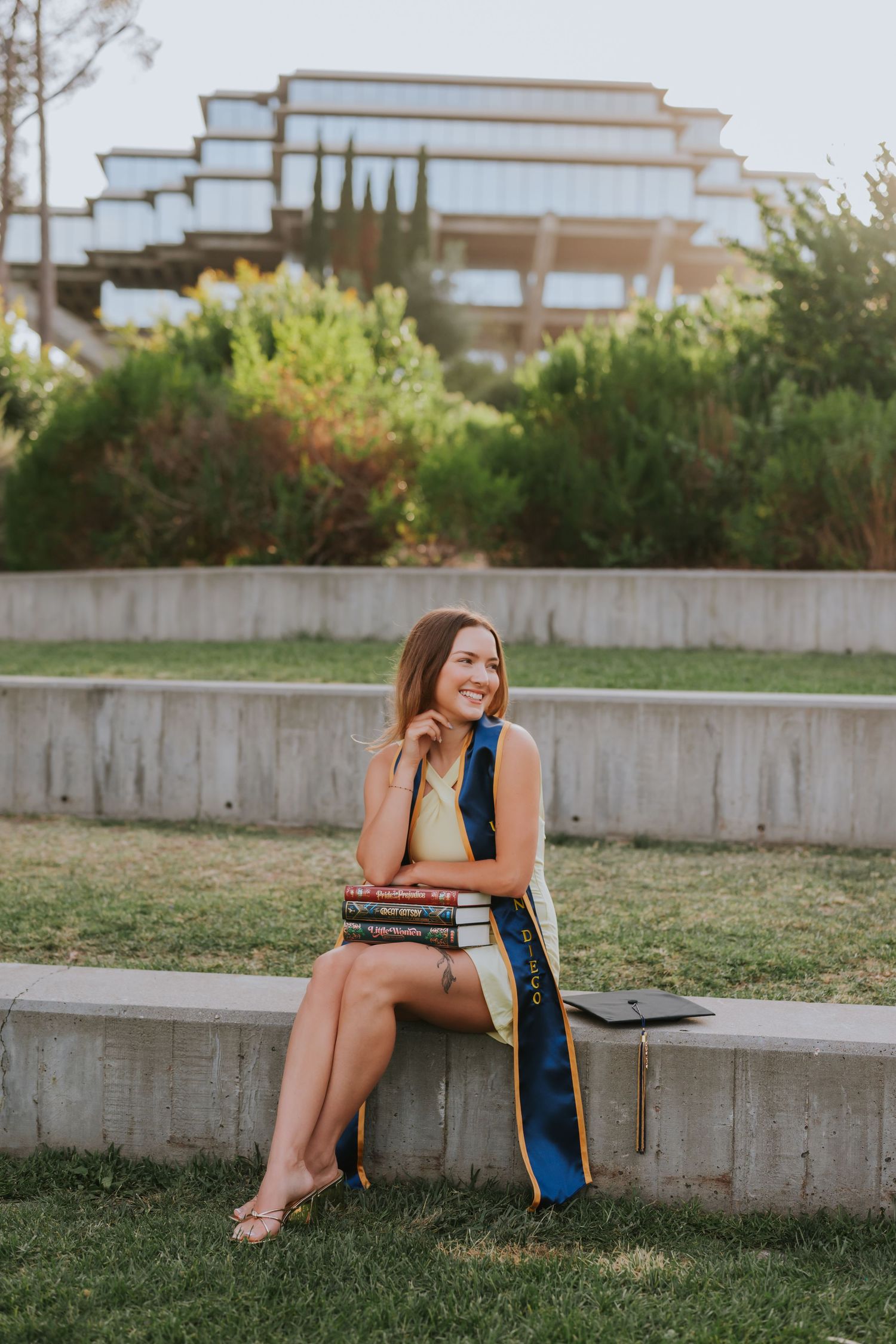 Smiling female graduate in blue sash sits outdoors with stacked books and cap, university building in background.