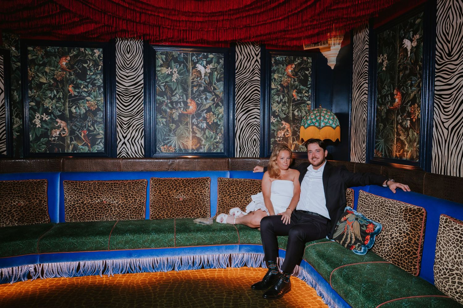 Bride and groom sitting together on a colorful leopard print sofa in an eclectic decorated room.