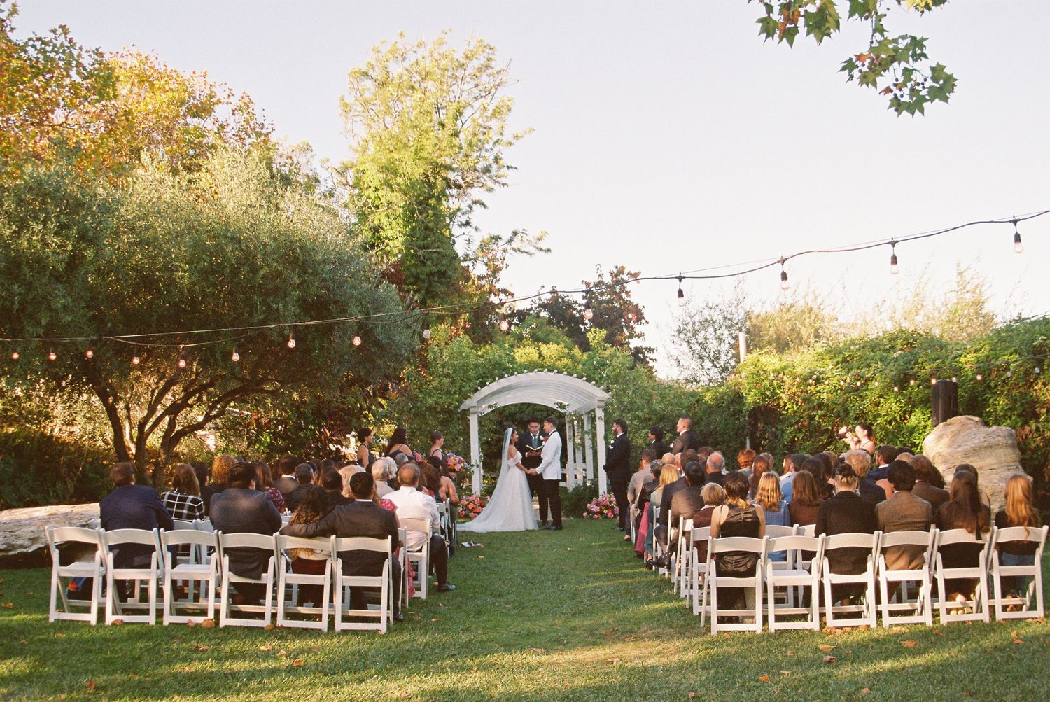 Outdoor garden wedding ceremony with guests seated on white chairs, string lights, and a floral arch altar.