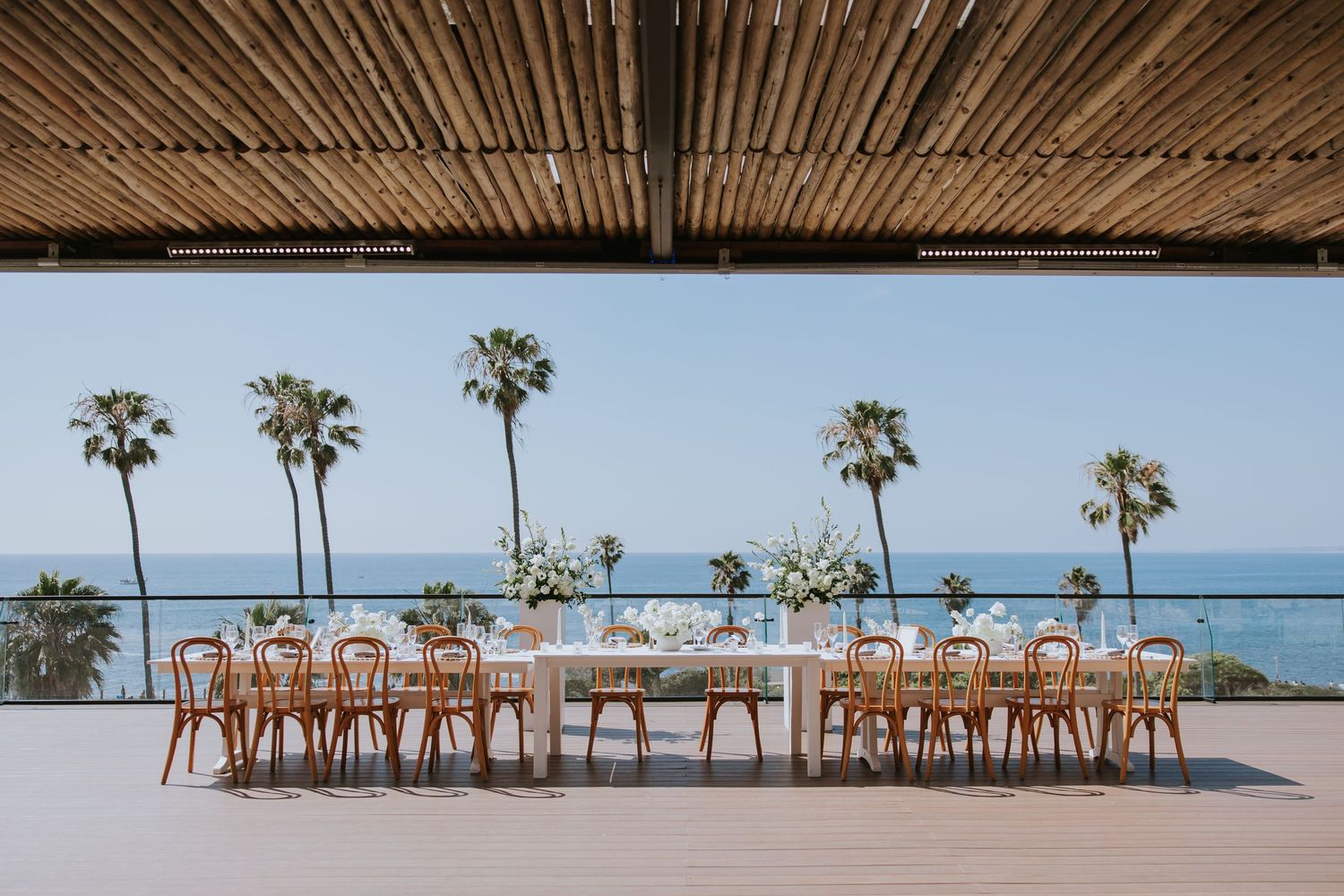 Elegant outdoor wedding reception setup with wooden chairs and tables along the beach with palm trees.