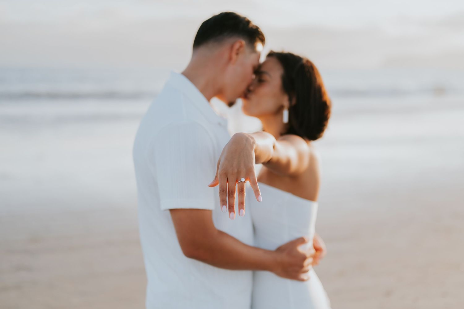A romantic couple in white embraces on a sandy beach during a sunset photoshoot.