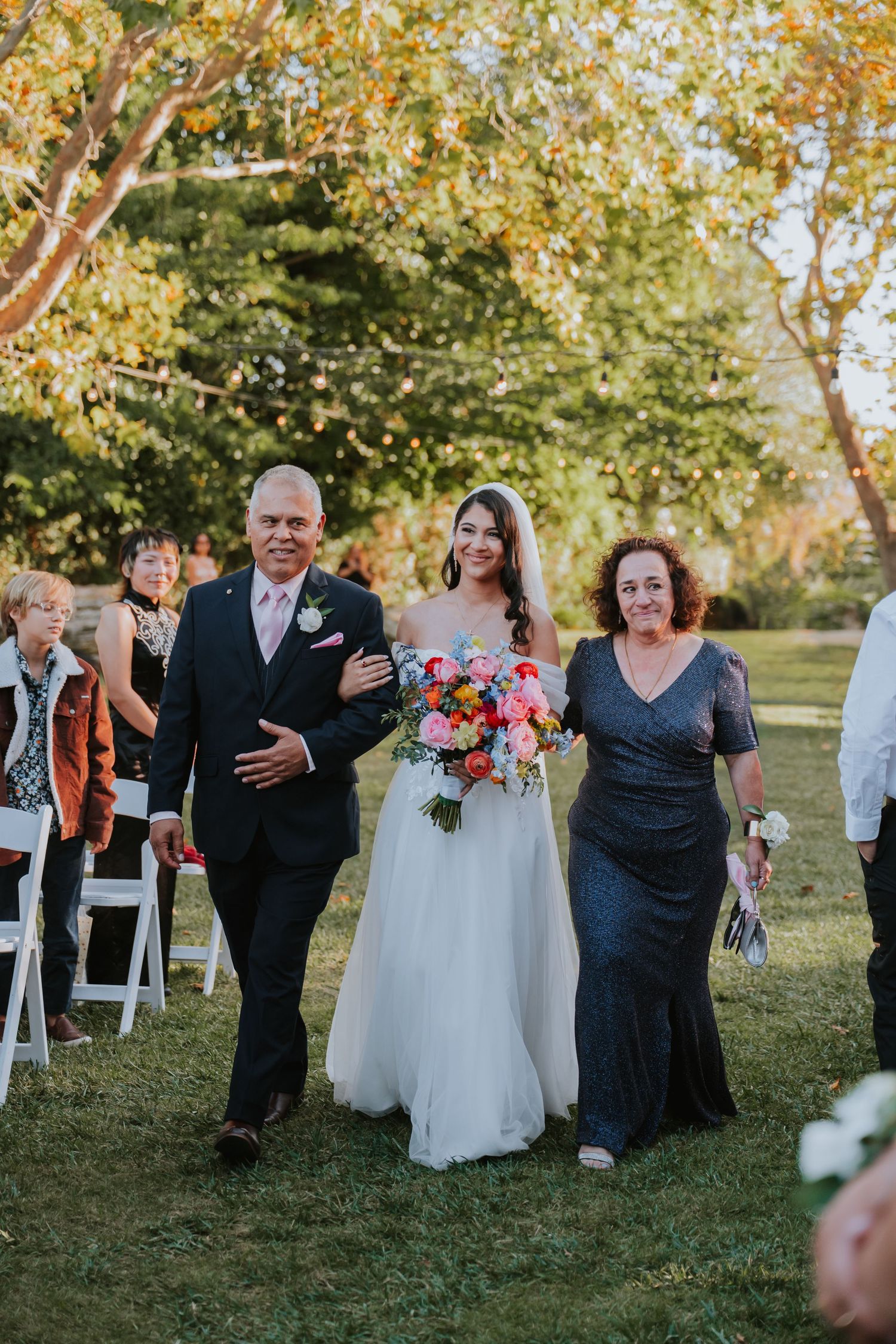 Wedding party walks together on grassy lawn during outdoor autumn ceremony with white chairs in background.