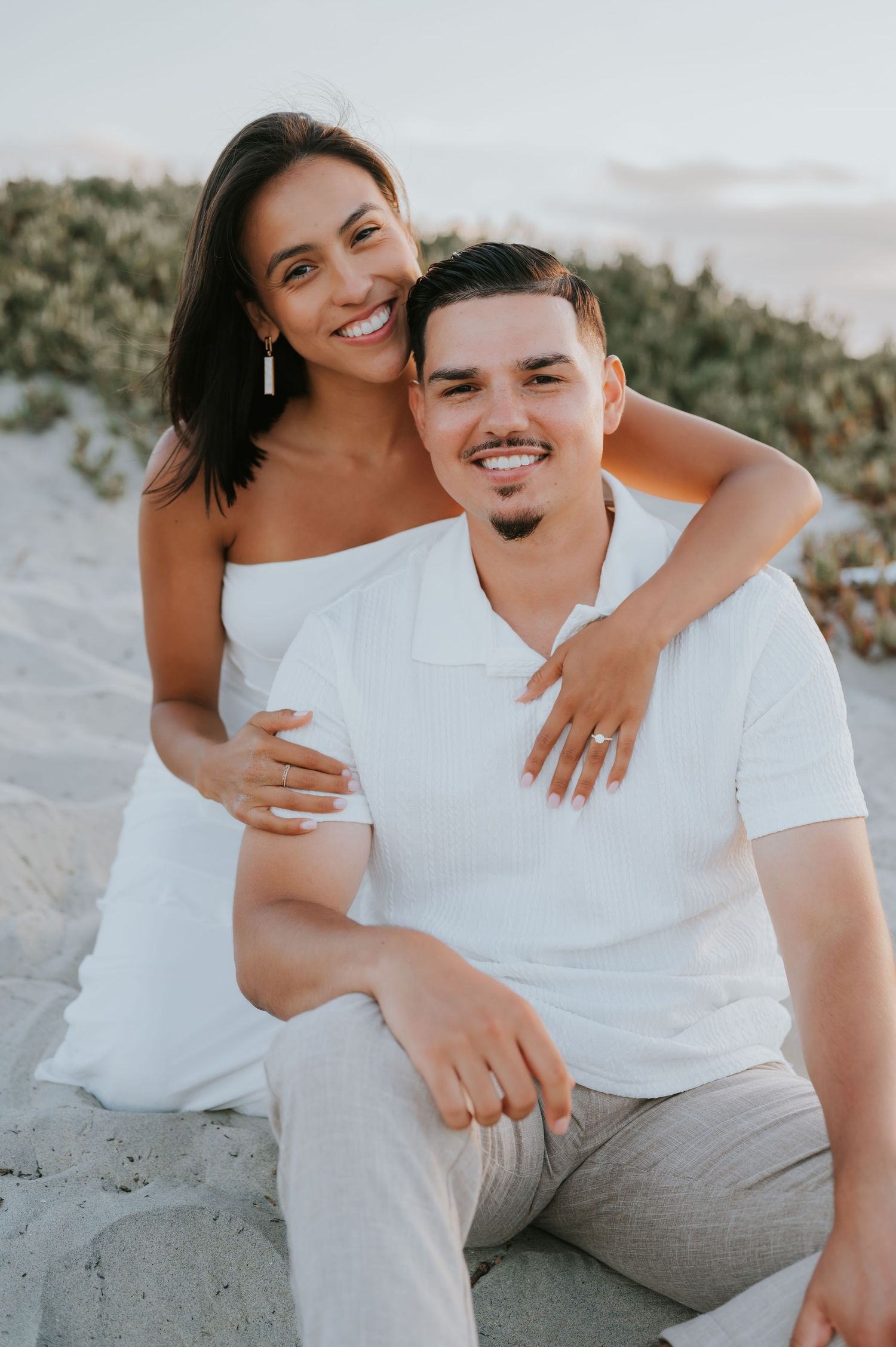 Couple wearing matching white outfits share intimate moments on beach during engagement session.