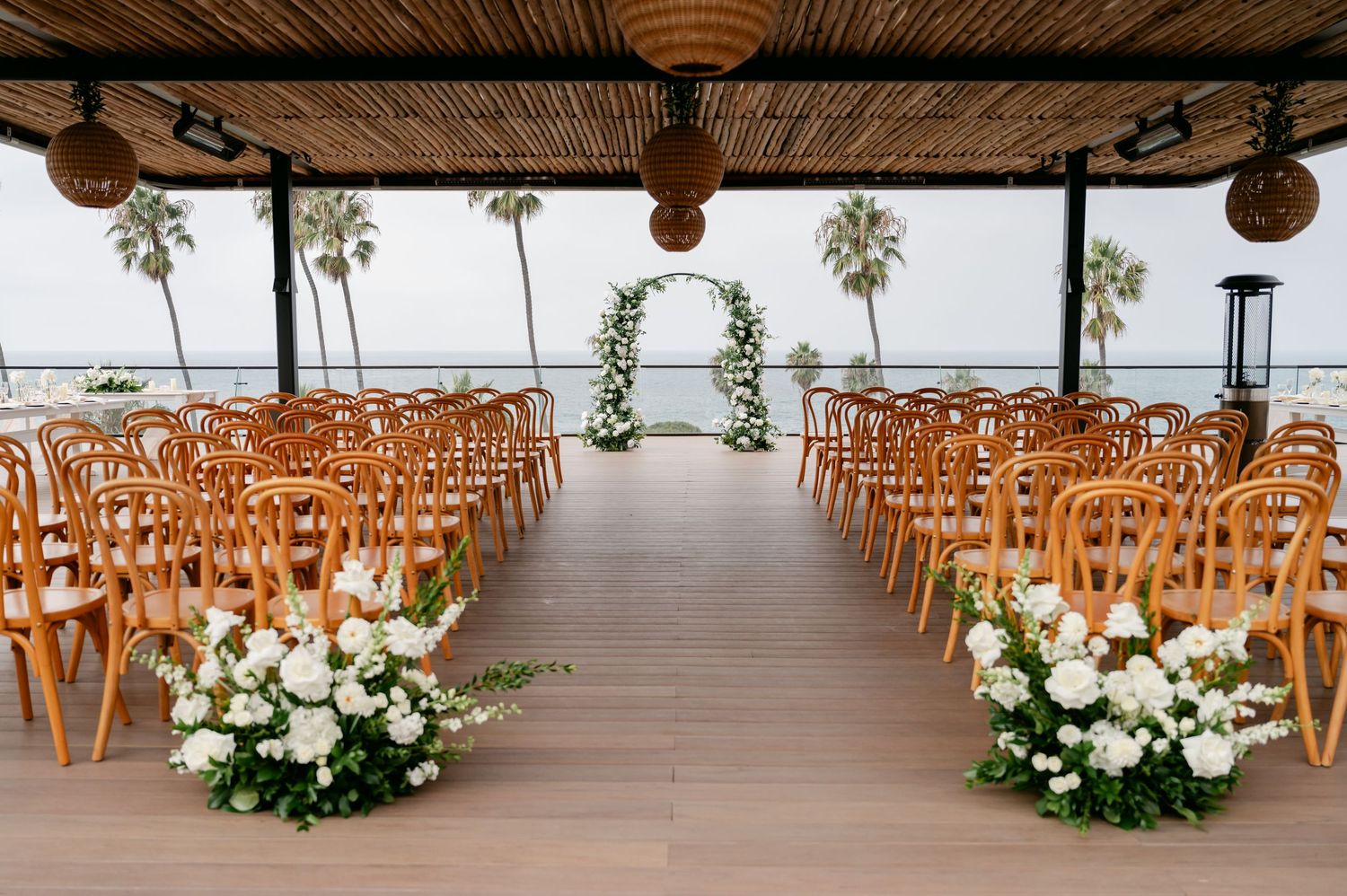 Covered oceanfront wedding venue with wooden chairs and white floral aisle decorations.