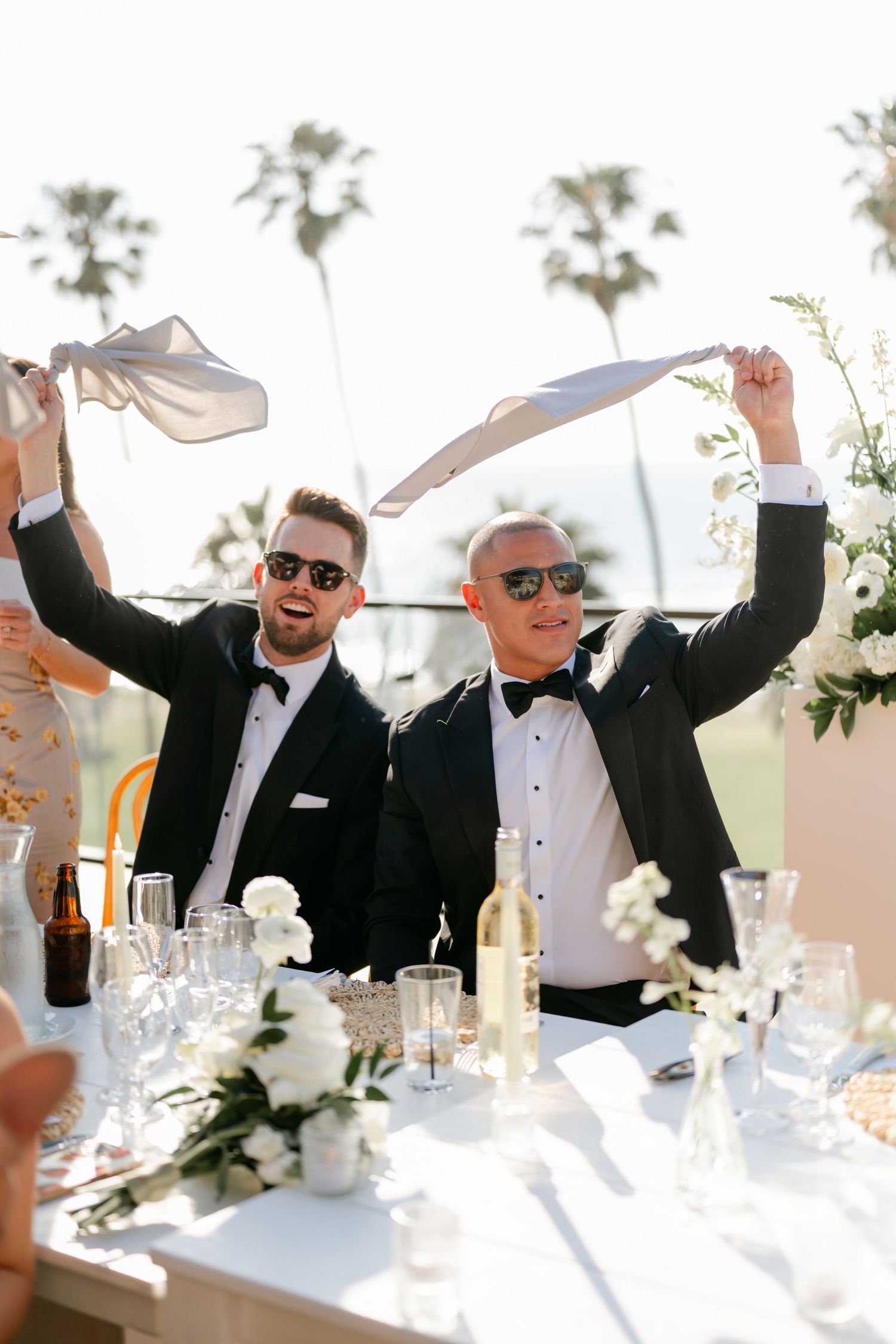 Wedding party celebrating at a beachside reception with white tablecloths and floral centerpieces.