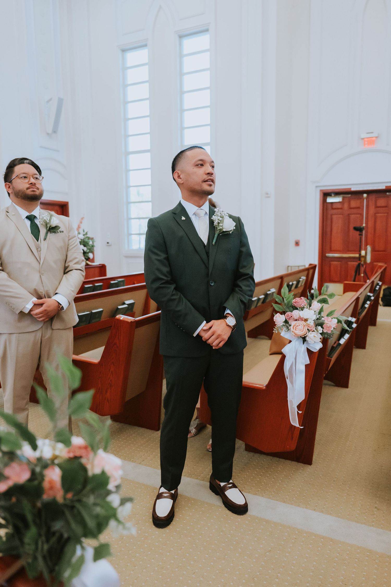 Groomsmen in dark and light suits stand in church pews during a wedding ceremony.