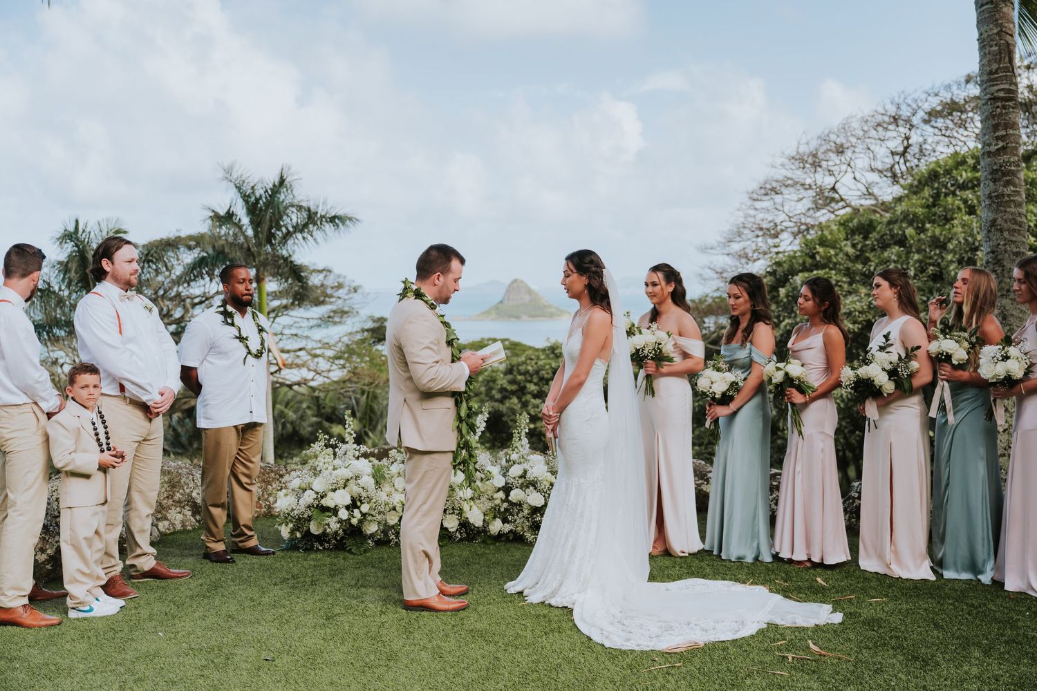 A wedding ceremony takes place on a hilltop overlooking the ocean with bridesmaids in pastel dresses and groomsmen in light suits.