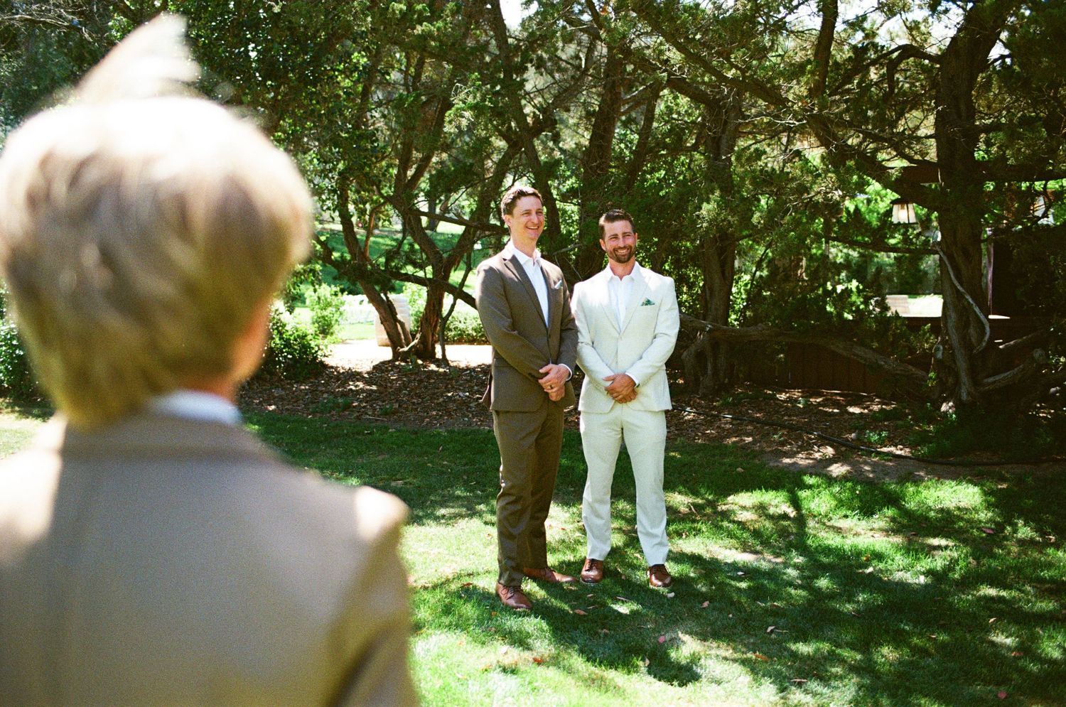 Two grooms stand together outdoors in a lush garden during an intimate same-sex wedding ceremony.