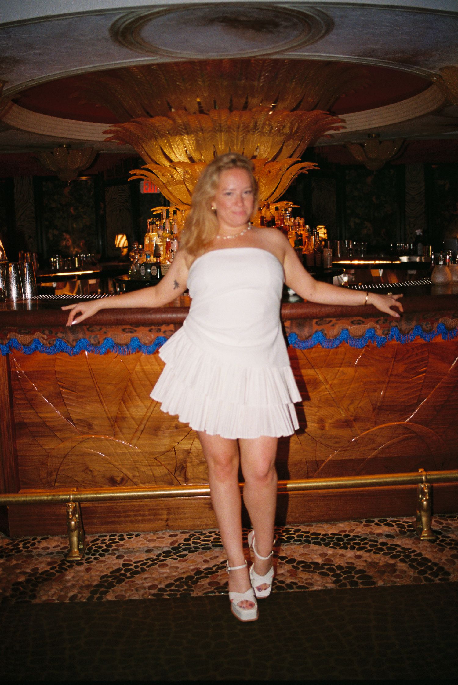 Woman posing confidently in white strapless mini dress and platform heels at a luxurious bar setting.