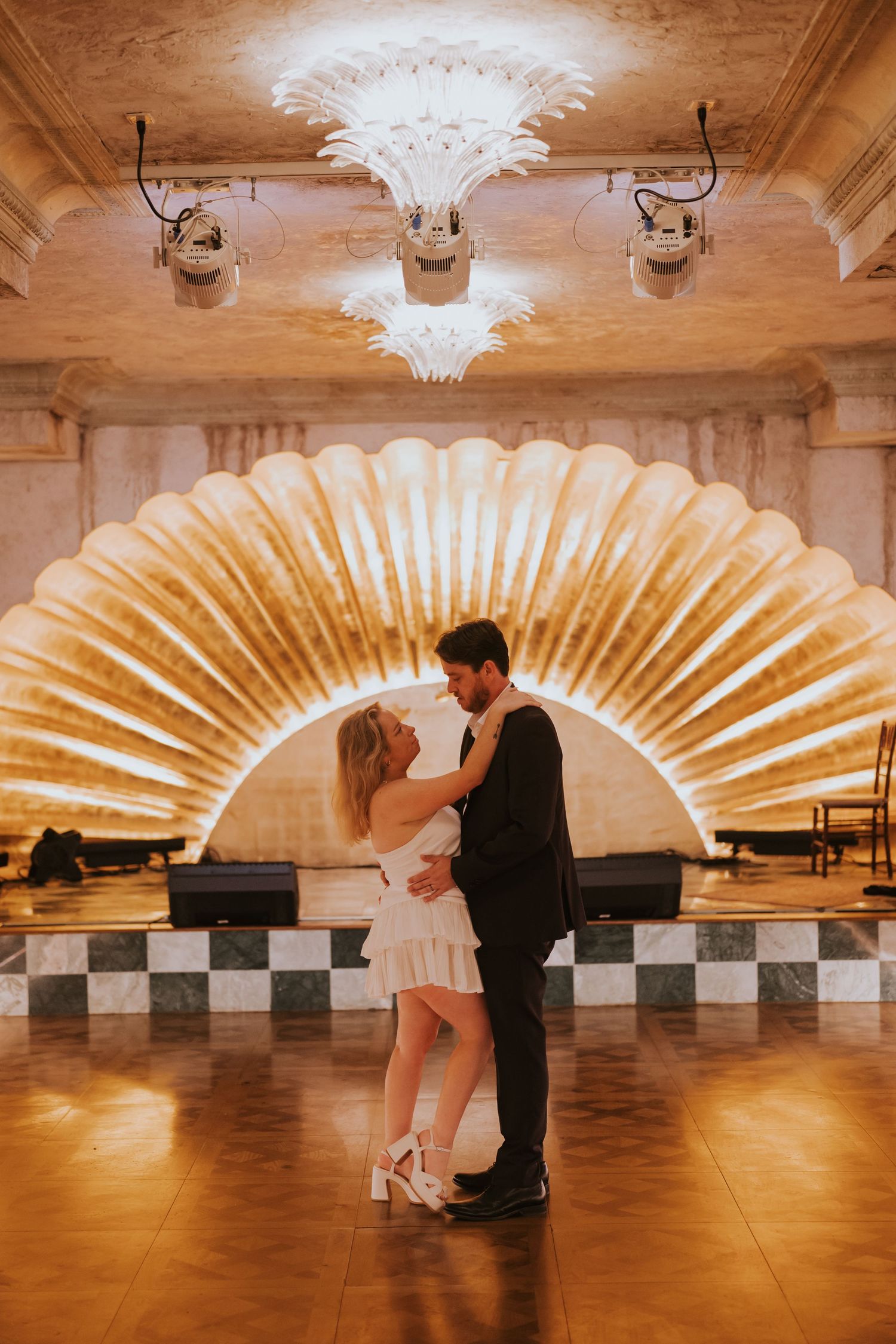 Bride and groom share a first dance beneath a glowing shell-shaped backdrop in an elegant ballroom.
