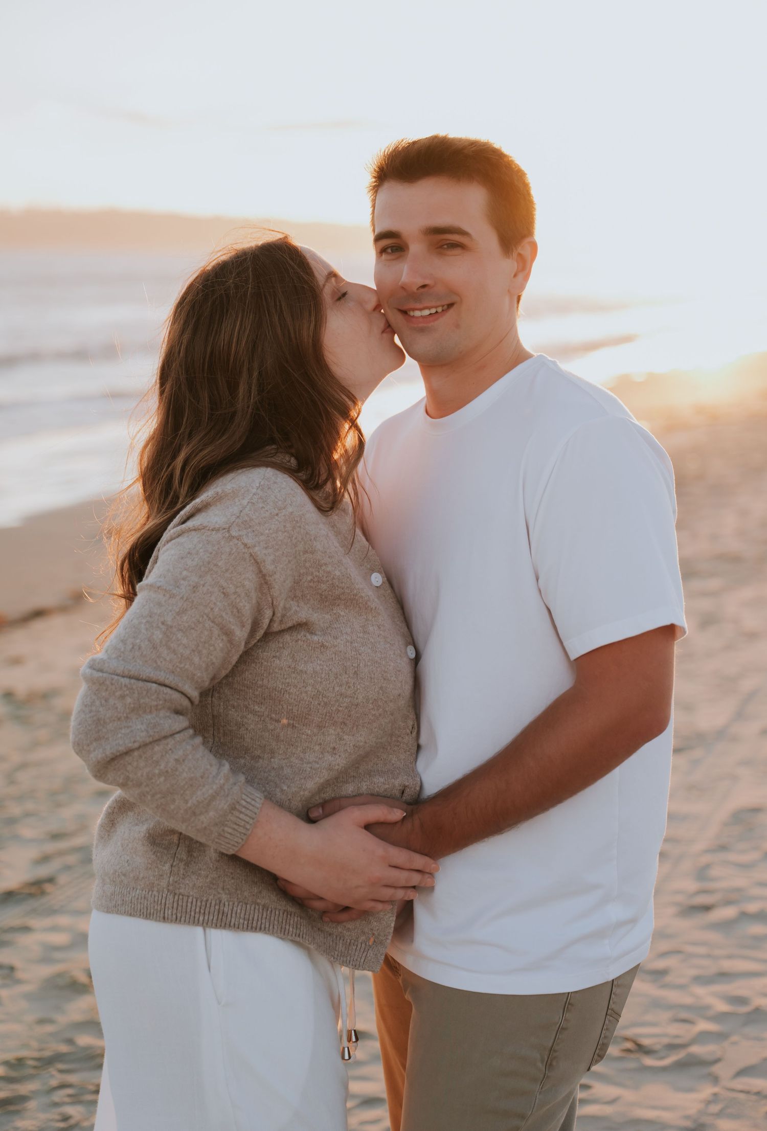 Couple sharing a tender moment on a sandy beach at golden hour, woman kissing man's cheek during maternity photo session.
