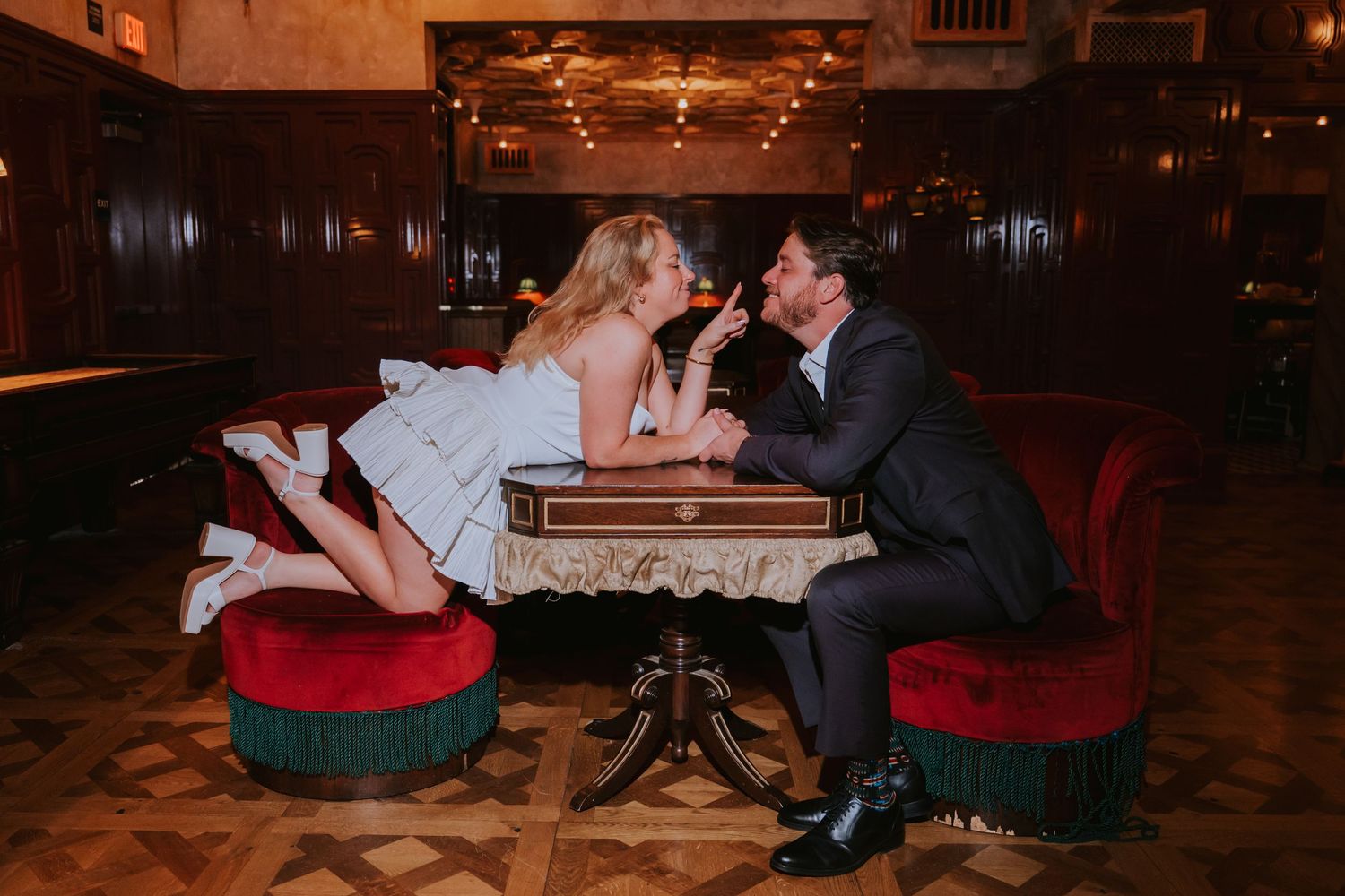 Playful bride leans across a small table to kiss groom in a warmly lit wood-paneled bar.