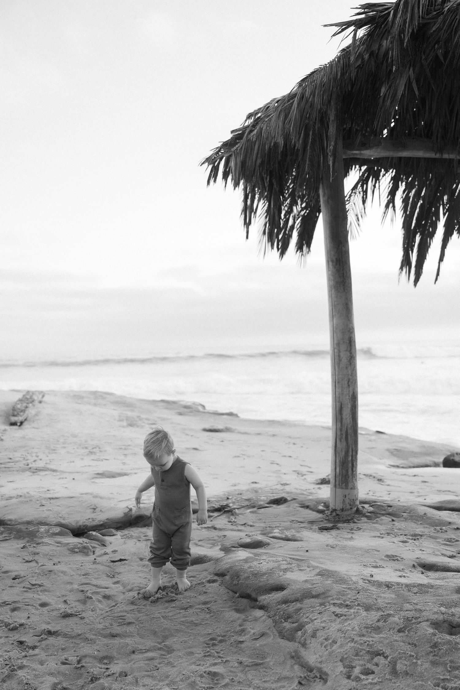 A toddler plays in the sand near a thatched palm umbrella on a beach with ocean waves in the background.