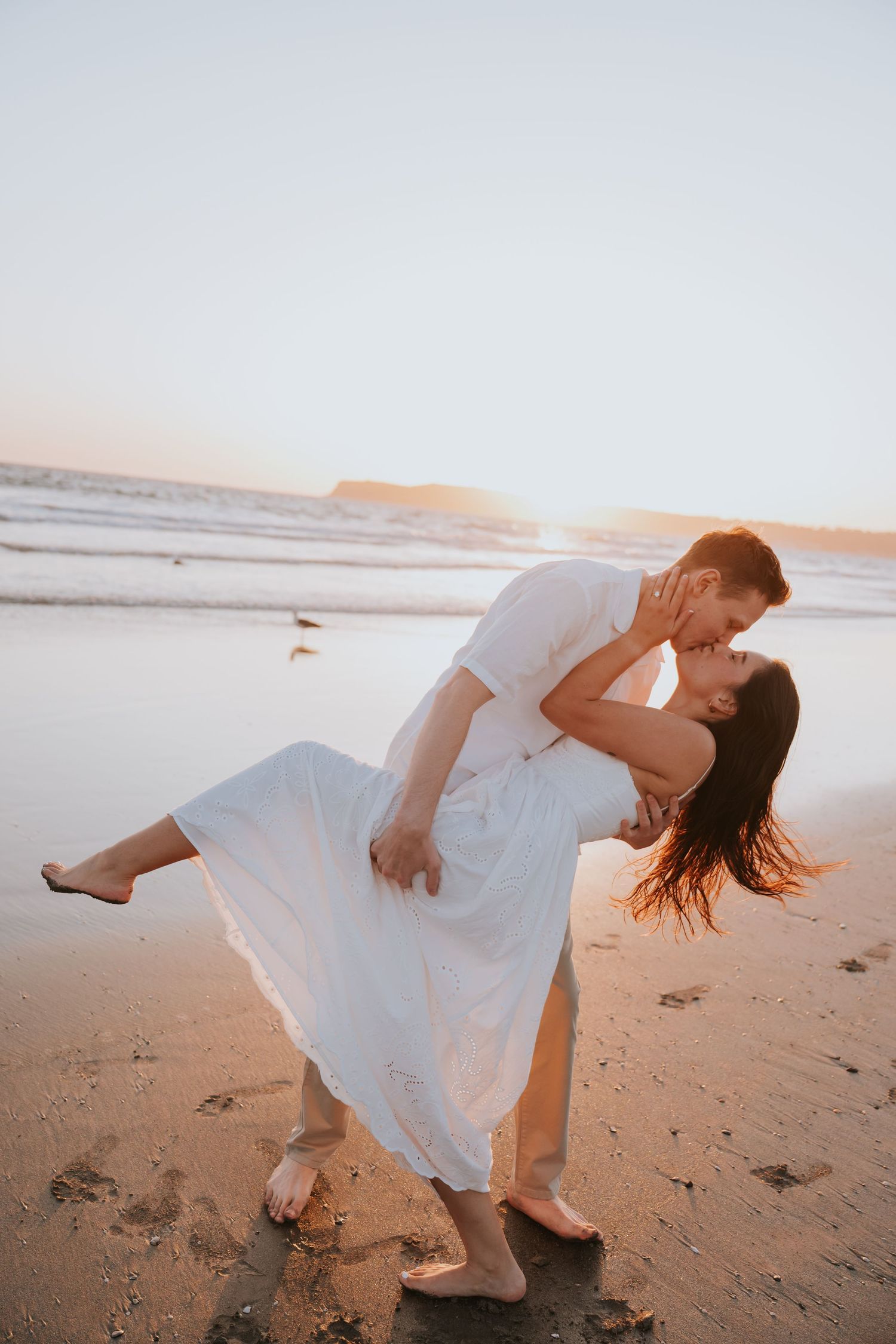 Couple sharing a romantic dip kiss on a sandy beach at golden sunset, woman in flowing white dress.