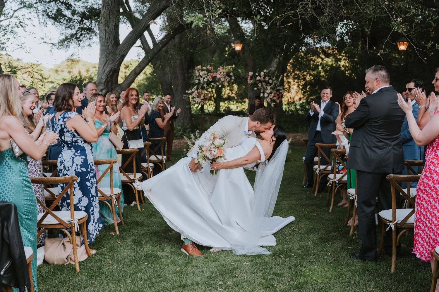 Bride and groom share a romantic dip kiss during outdoor wedding ceremony while guests applaud on a sunny day.