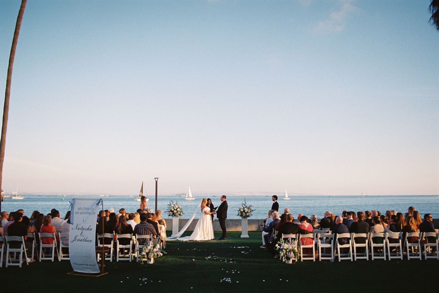 Outdoor beach wedding ceremony with guests seated in white chairs facing couple at altar with ocean view.