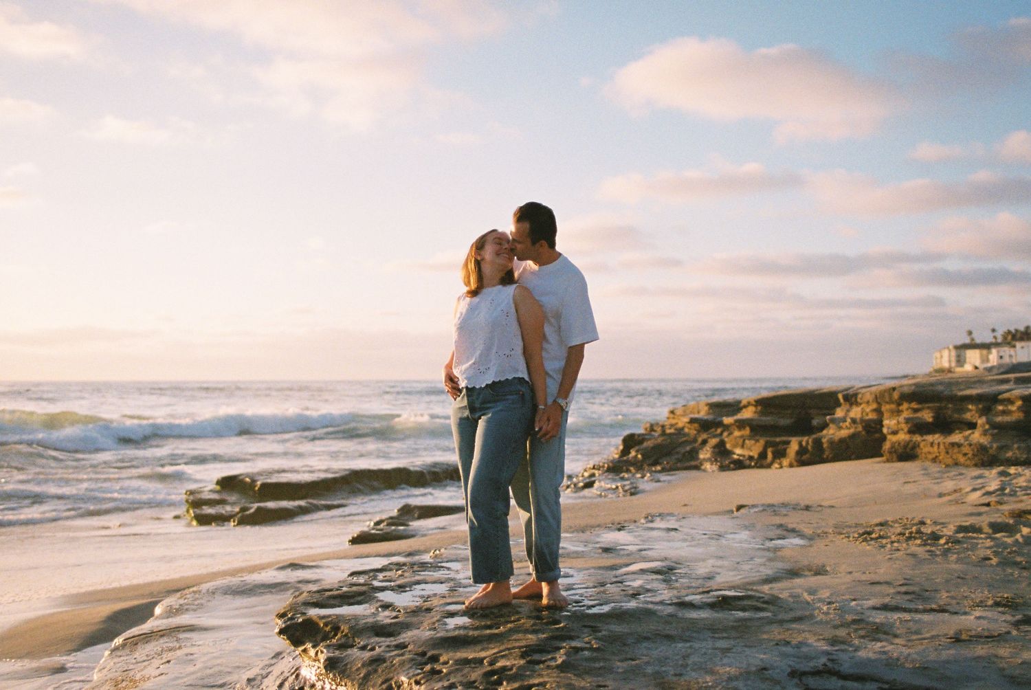 Couple sharing a romantic kiss on a rocky beach at sunset with ocean waves and pastel sky in the background.