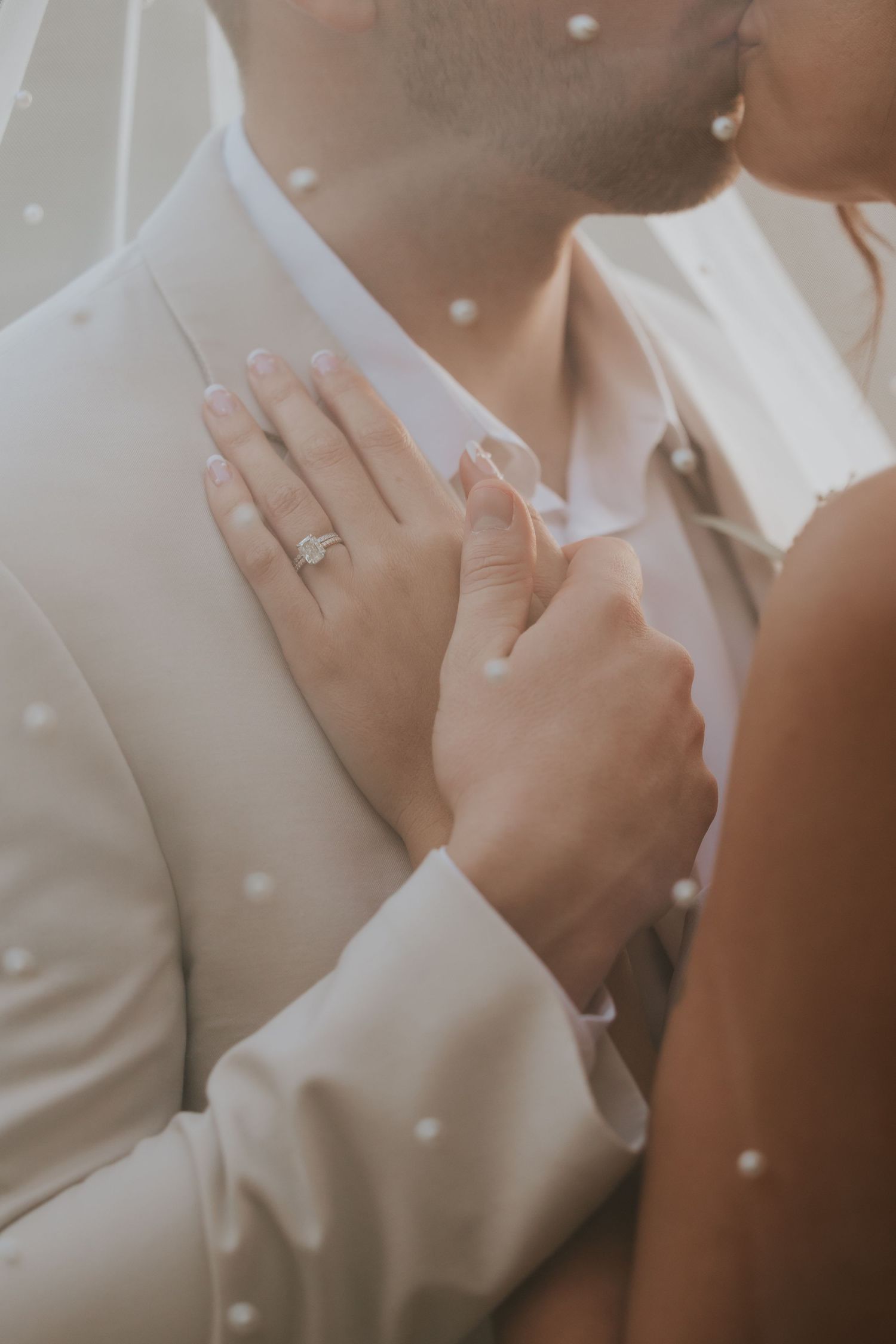 A close-up view of hands wearing an engagement ring against a soft white background with bokeh effects.