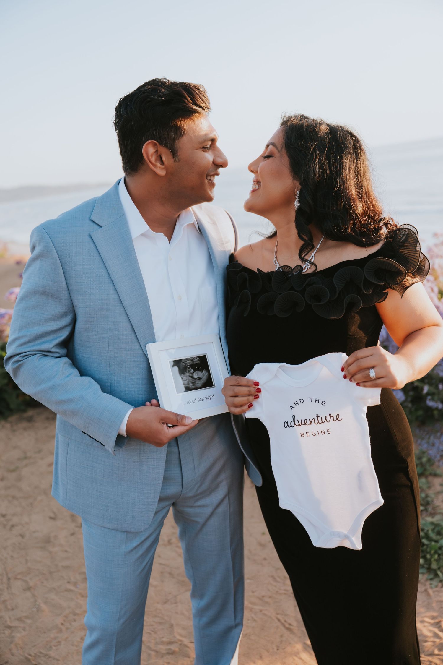 Expecting couple smiling at each other outdoors, holding ultrasound photo and baby onesie reading 'and the adventure begins'.