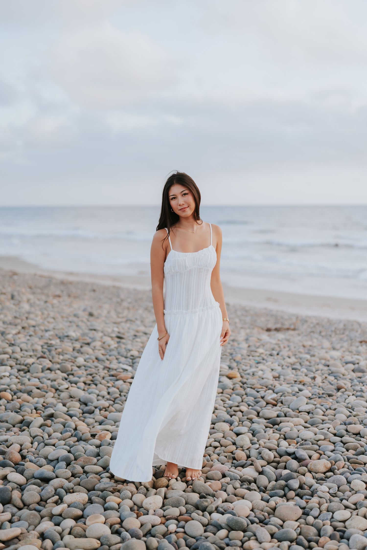 Woman in white maxi dress standing on rocky beach with ocean waves in background.