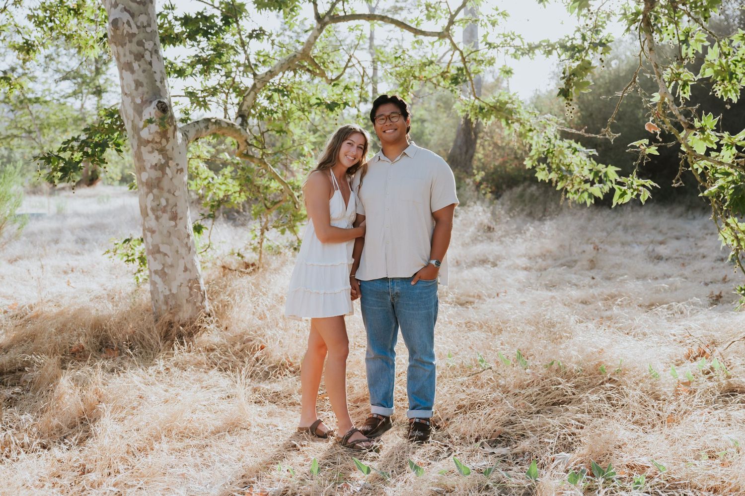 Couple posing together outdoors under a tree in a sunny, dry landscape, woman in white dress, man in jeans.