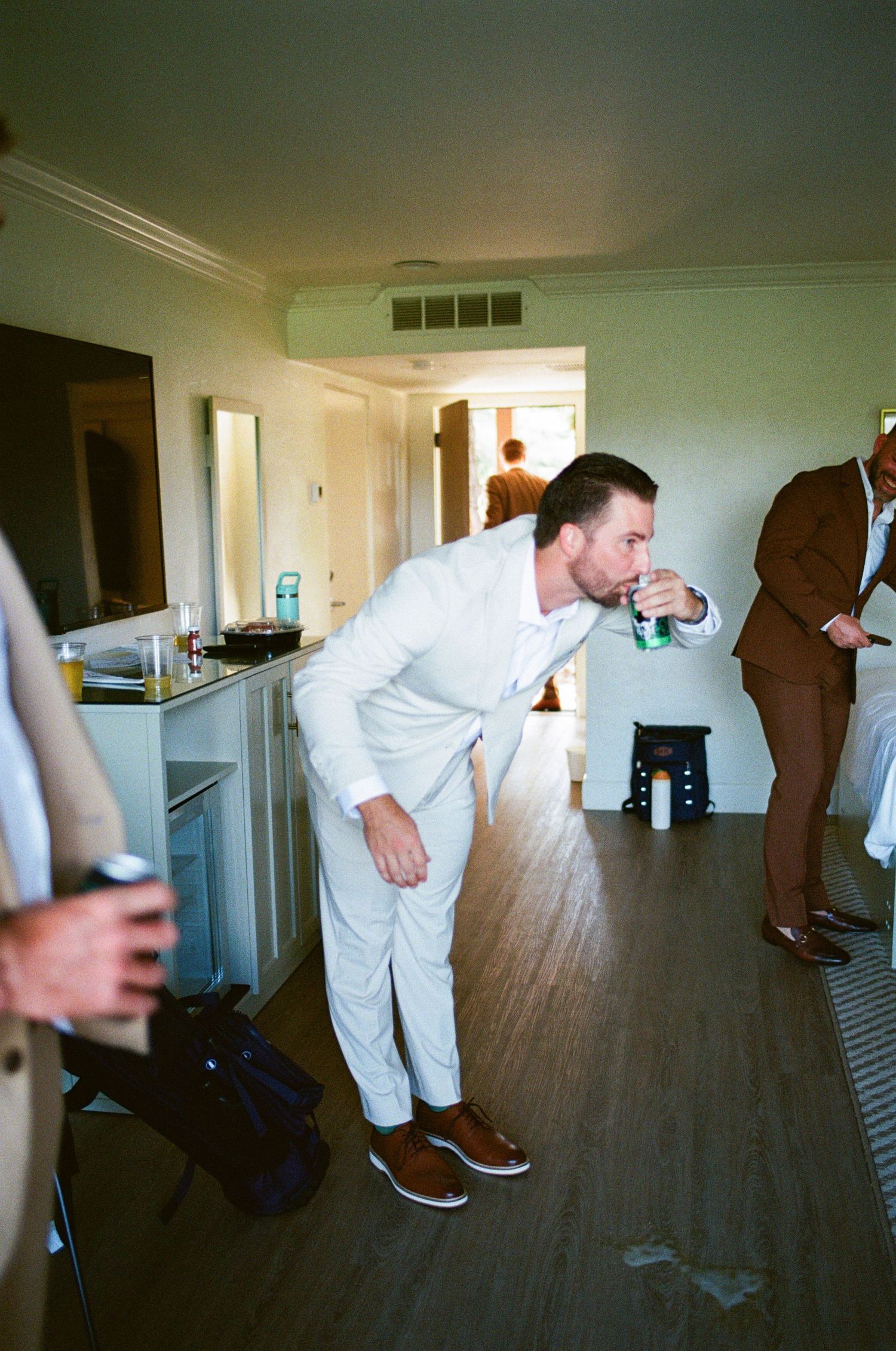 Groom in white suit drinking beer while getting ready with groomsmen.