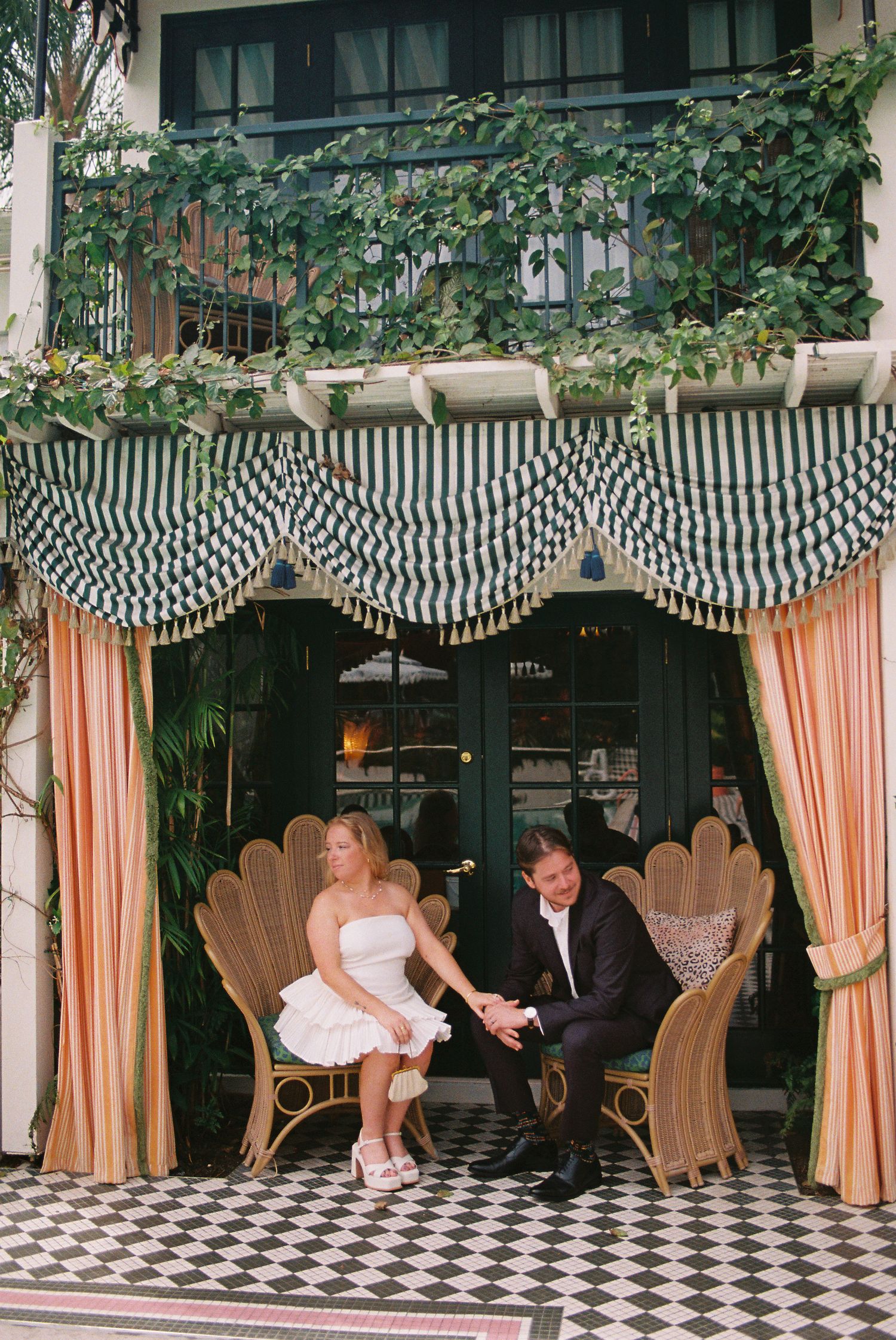 Couple sitting in ornate wicker chairs under striped awning with peach curtains at elegant venue.