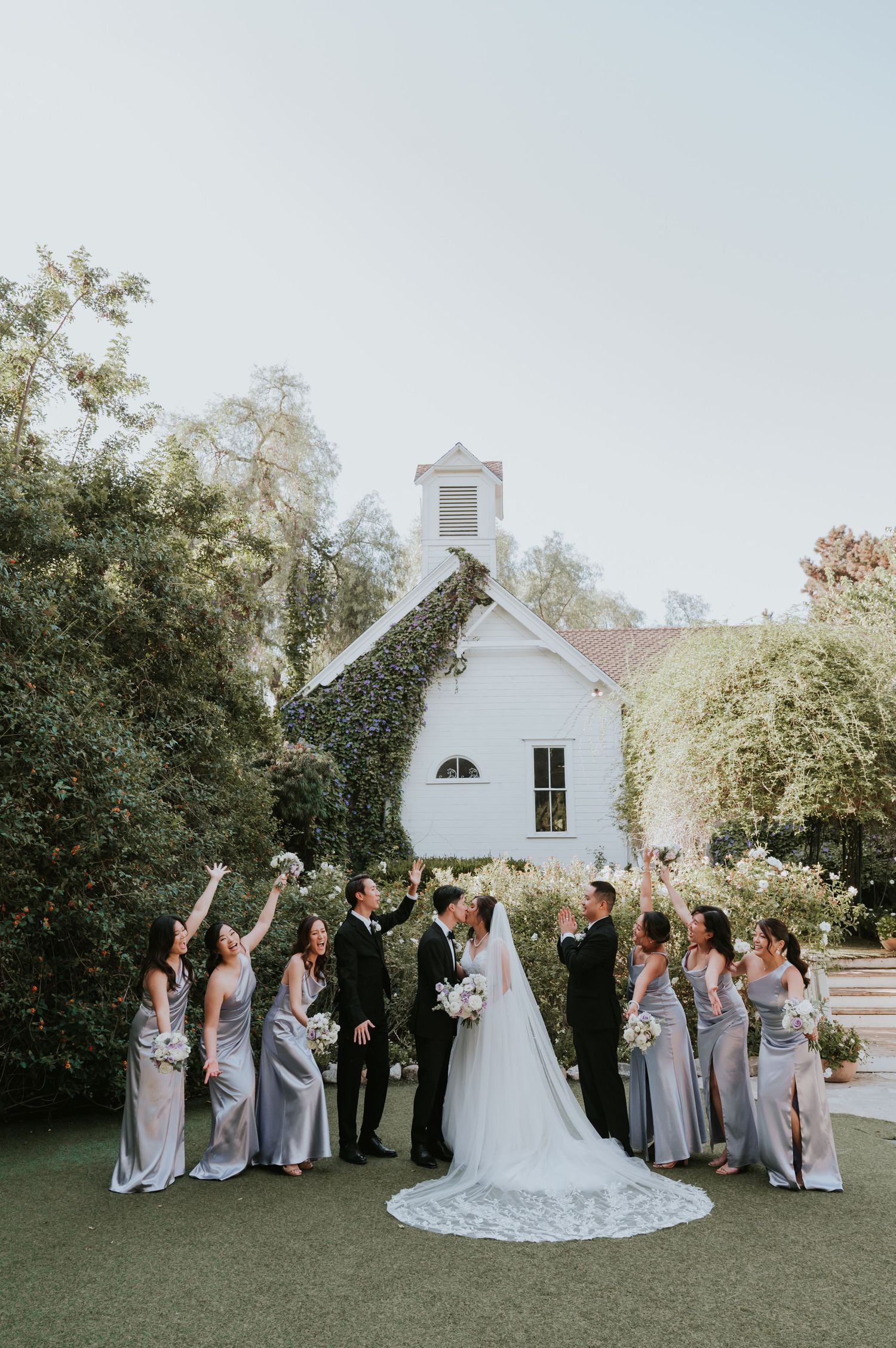 Wedding party in grey dresses and black tuxedos poses in front of white chapel during outdoor ceremony.