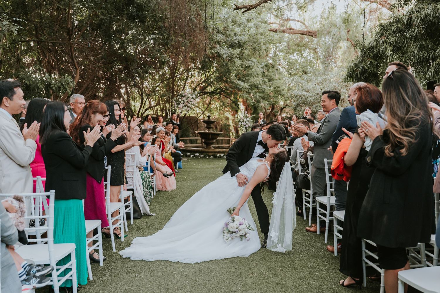 Outdoor wedding ceremony with guests seated in white chairs on garden lawn.
