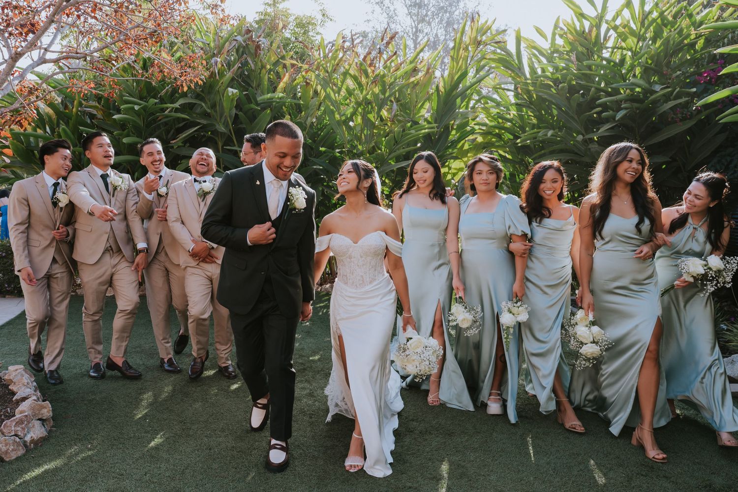 Wedding party in light colored dresses walks together through a garden setting during an outdoor celebration.