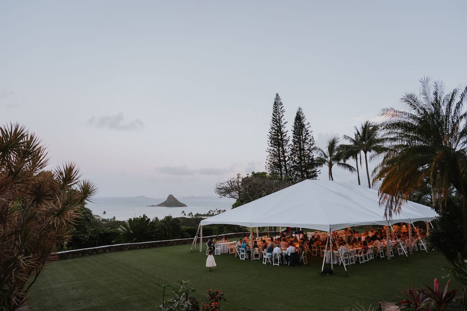 Panoramic view of an outdoor wedding reception setup with white tent against ocean and mountain scenery.