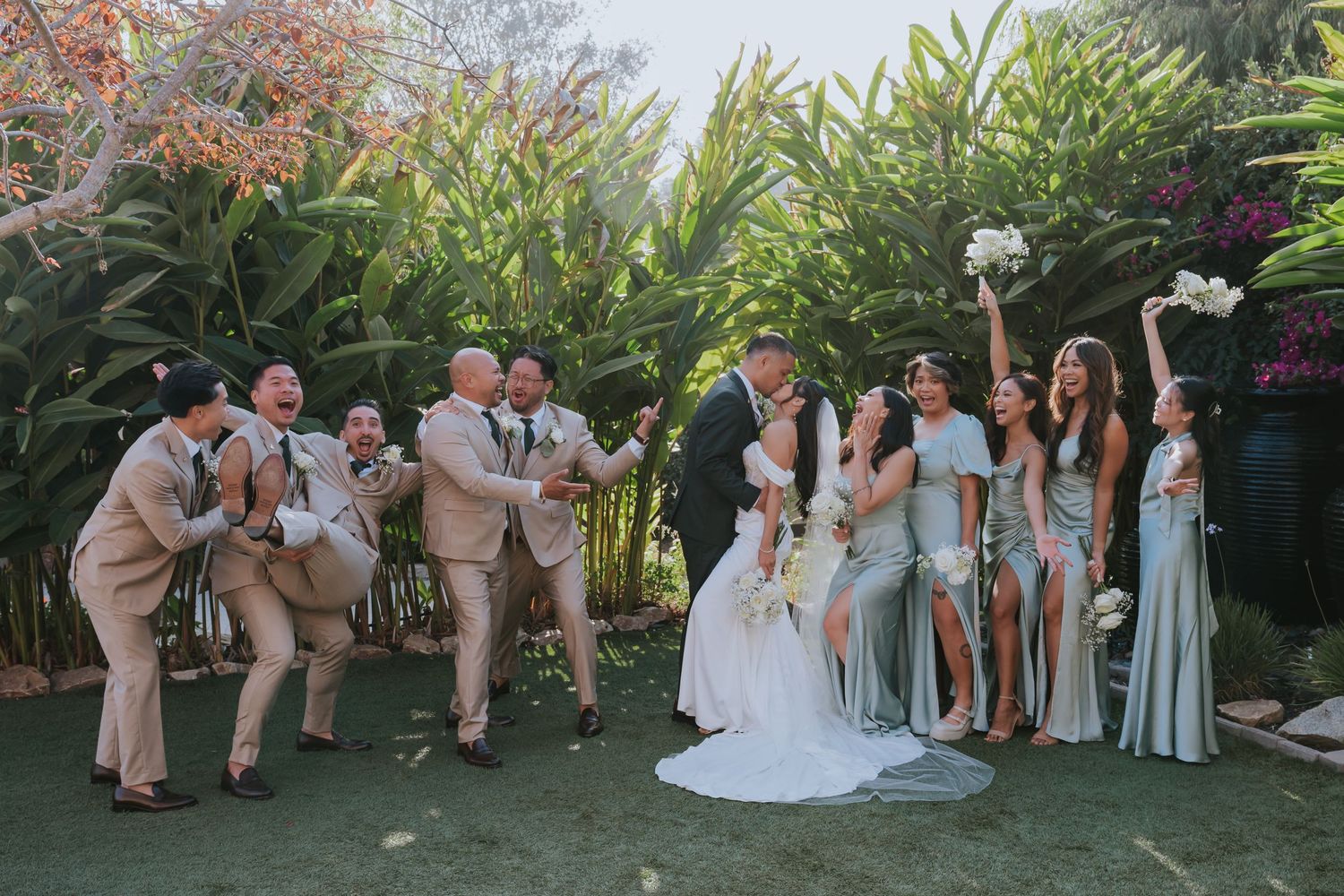 Wedding party in light blue dresses and tan suits poses together in a lush garden setting for formal photos.