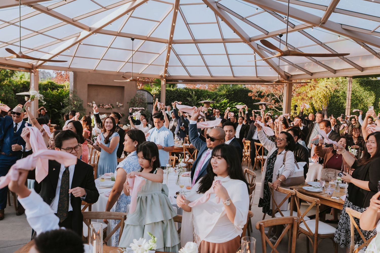Wedding guests dance and celebrate on an outdoor dance floor under a clear tent during a lively reception party.