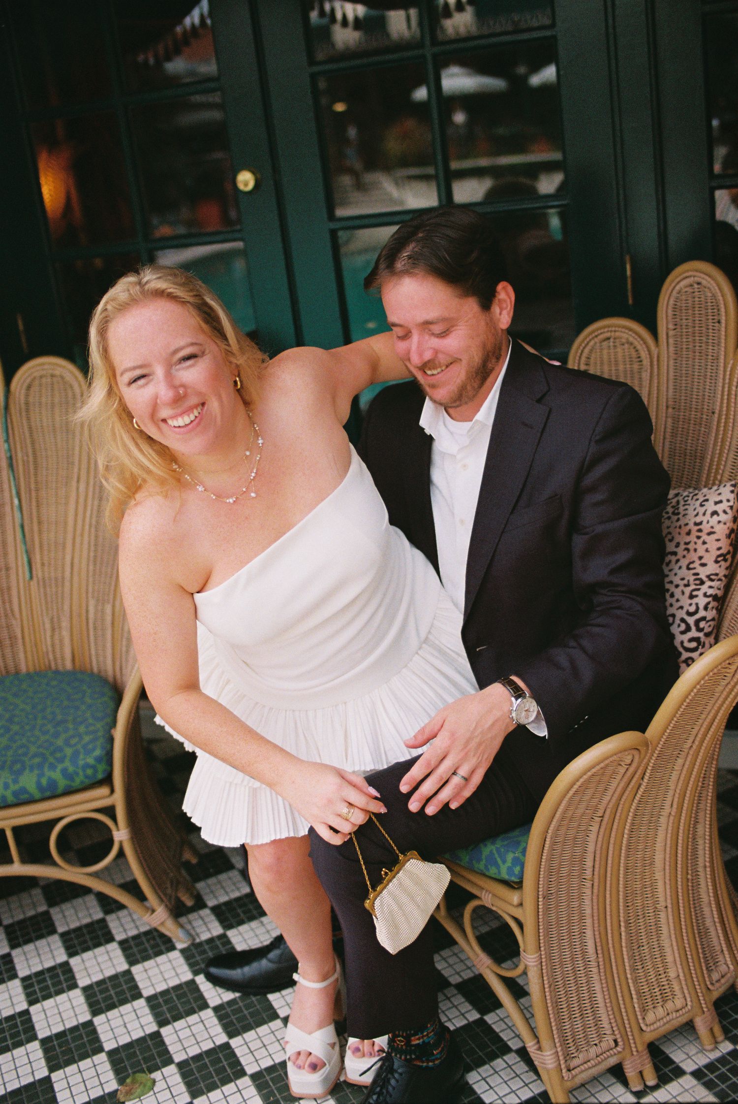 Smiling bride in white mini dress sits with groom in black suit on wicker chairs at an outdoor cafe.