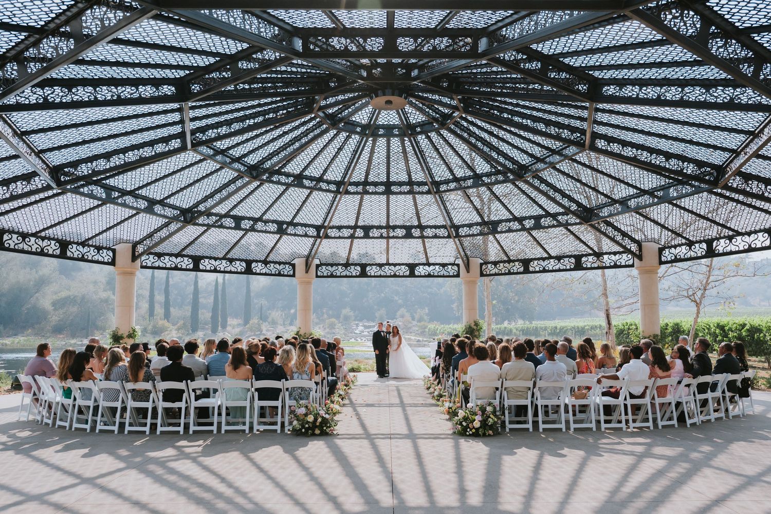 Elegant outdoor wedding ceremony under ornate glass dome gazebo with white chairs and floral arrangements.
