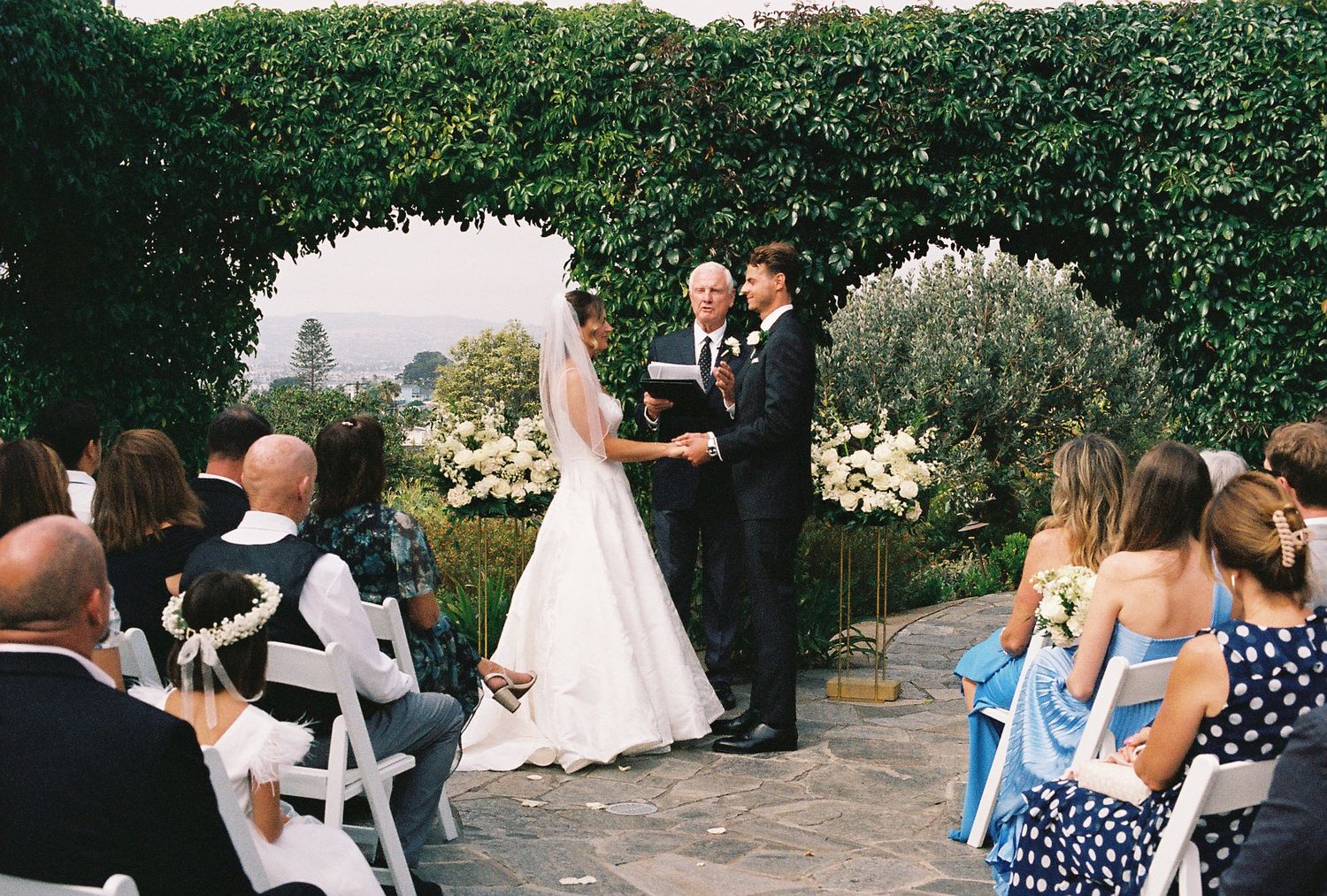 Outdoor wedding ceremony with bride and groom exchanging vows before officiant, surrounded by lush greenery and white floral arrangements.