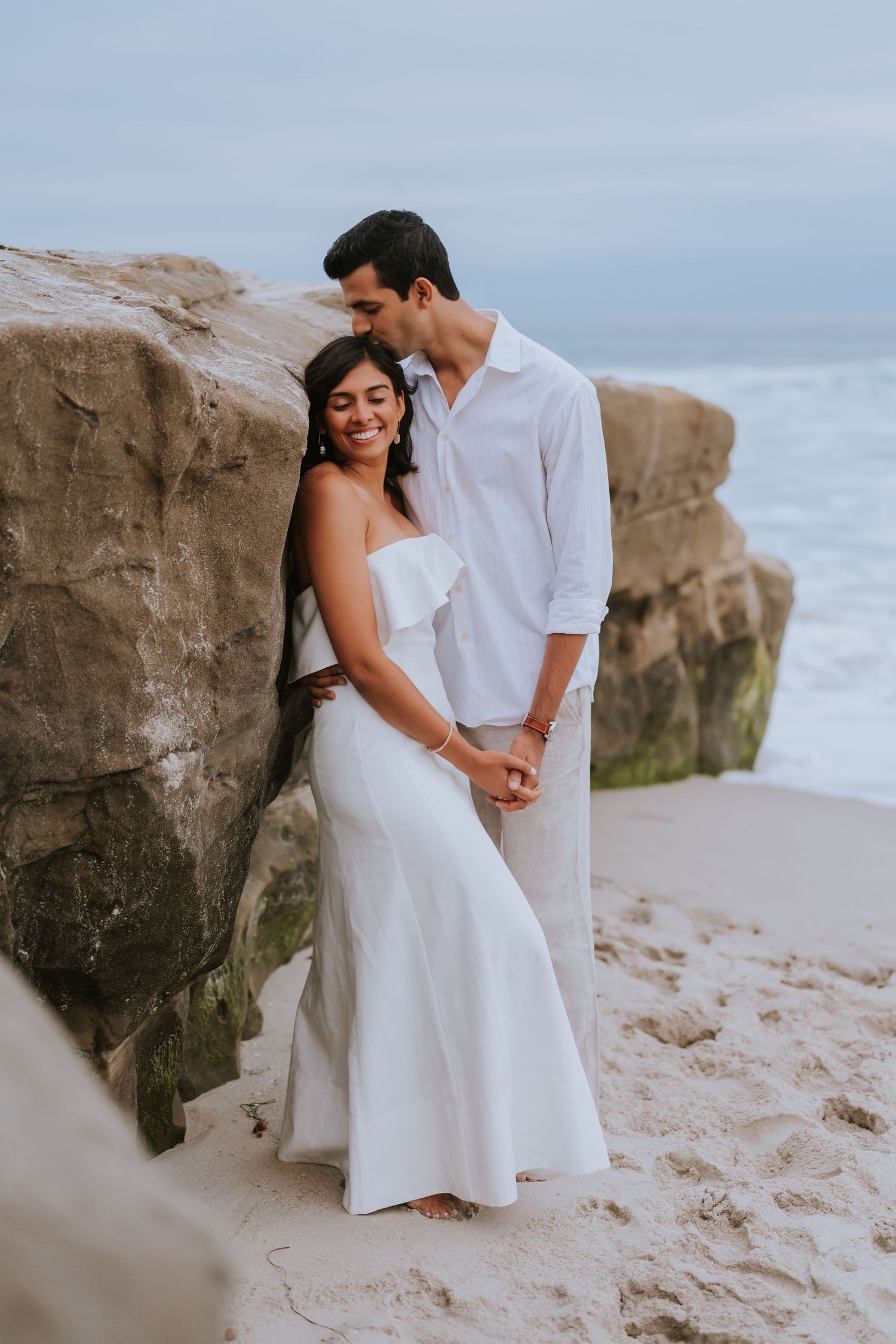 Romantic beach photoshoot with couple in white attire against rocky coastal backdrop.
