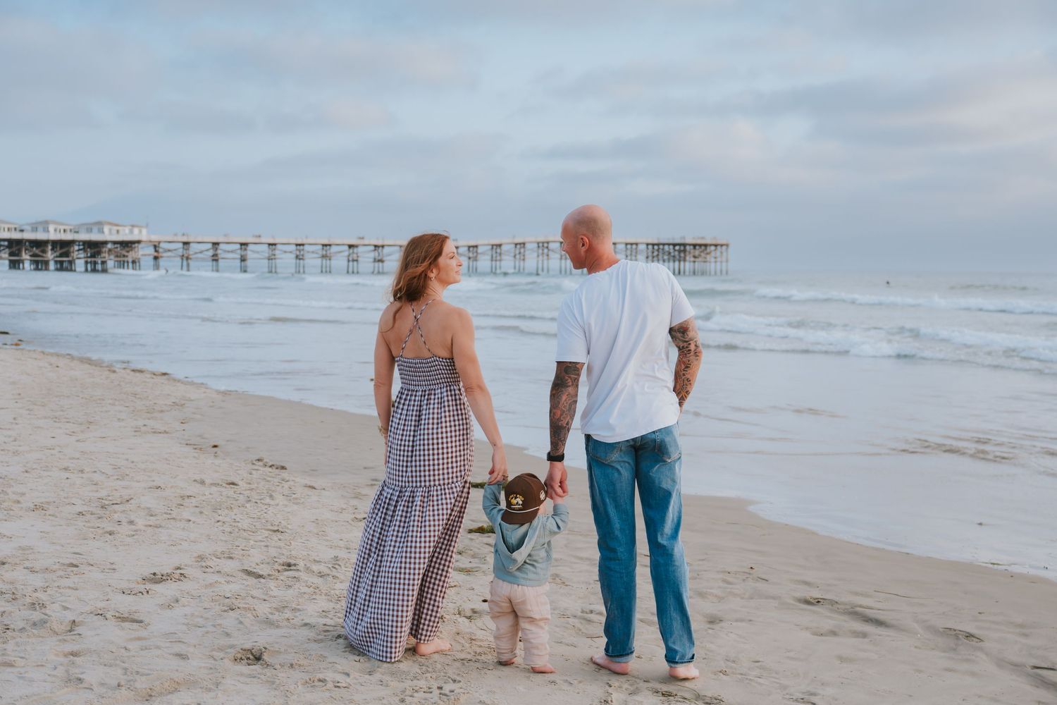 Family of three holding hands on a sandy beach at sunset with a pier in the background.