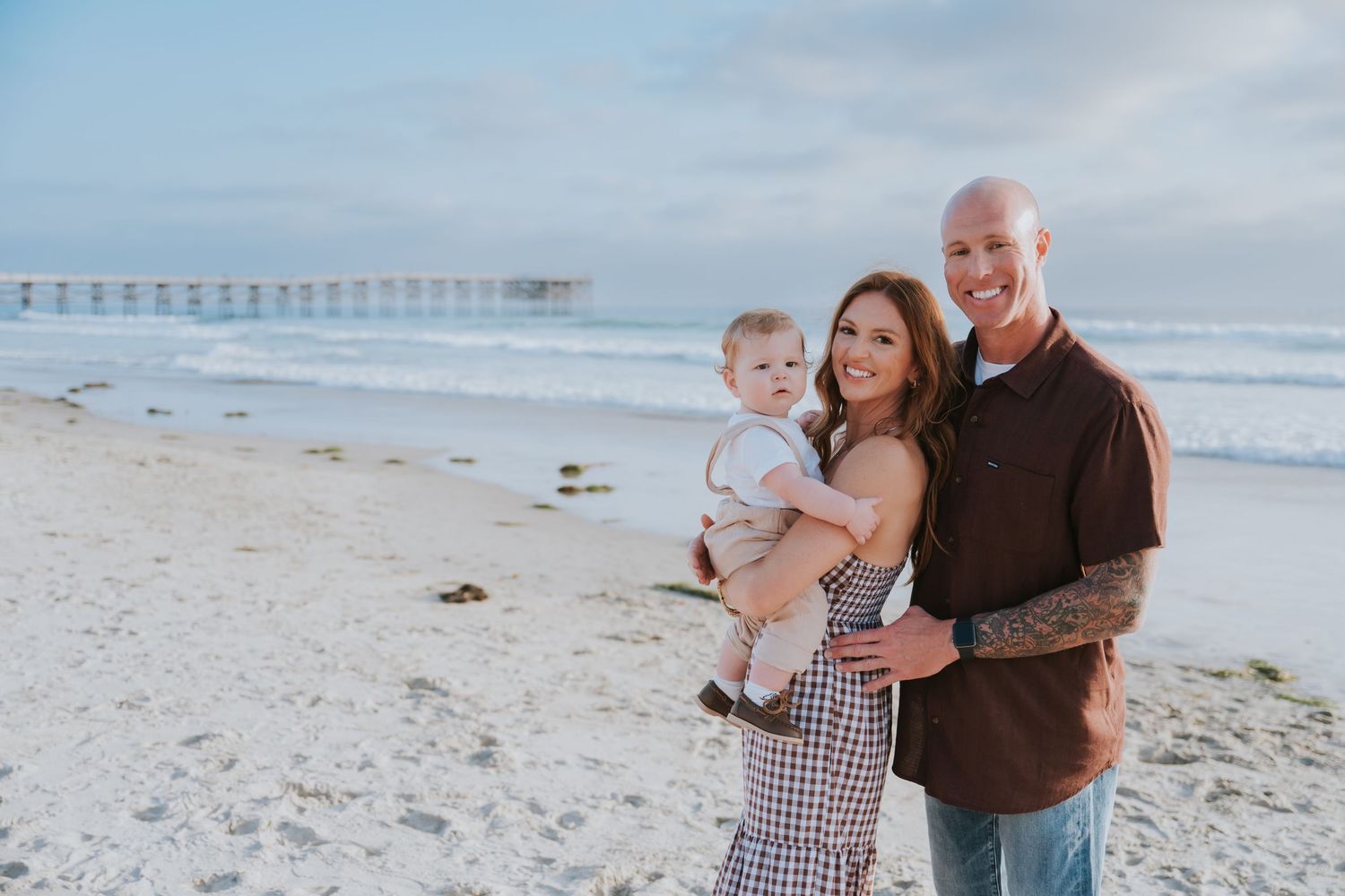 Happy family of three poses on a sandy beach near a pier, parents smiling while holding their baby boy.